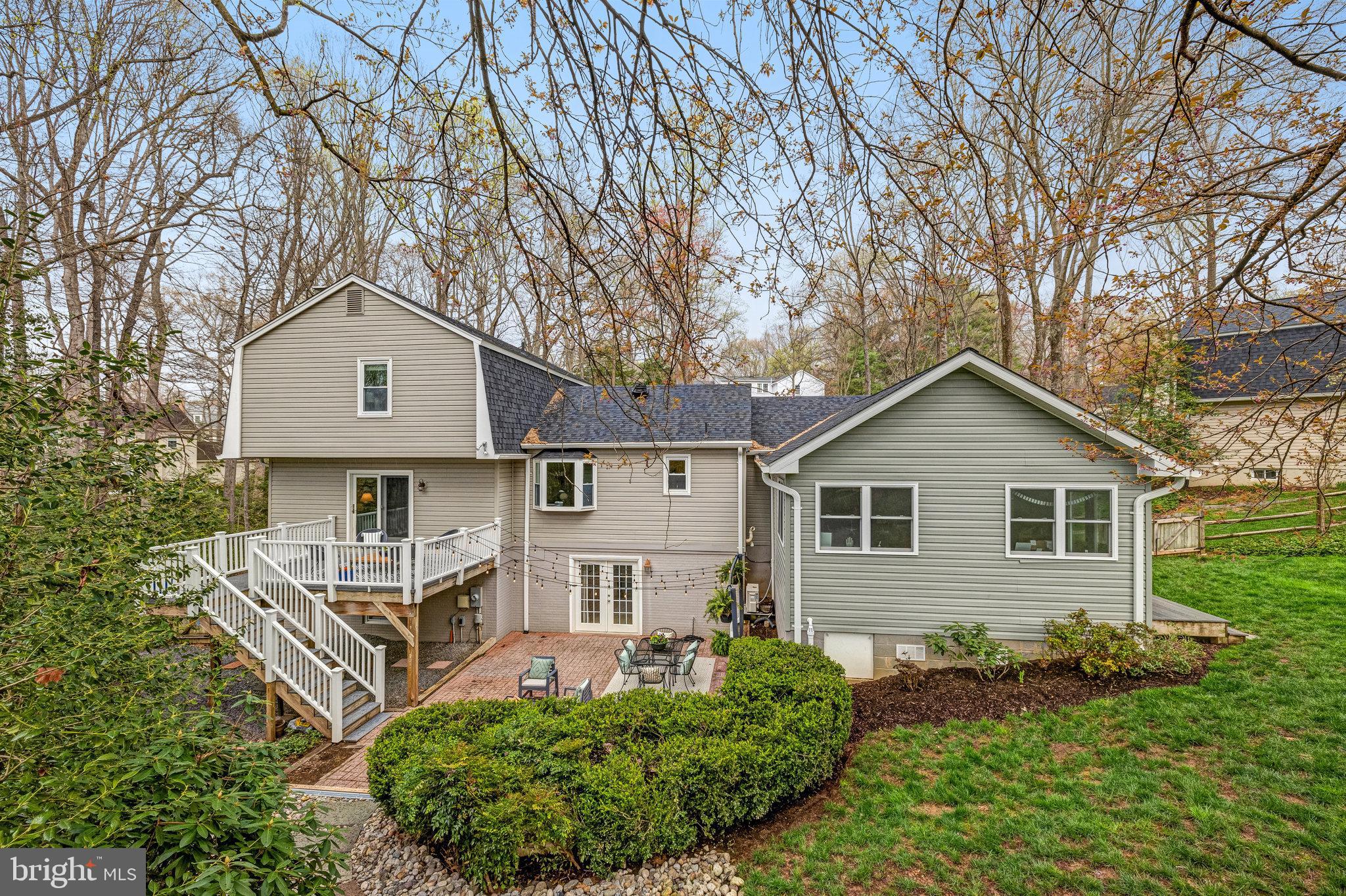 Home’s rear exterior with a raised deck, brick patio, and mature trees.