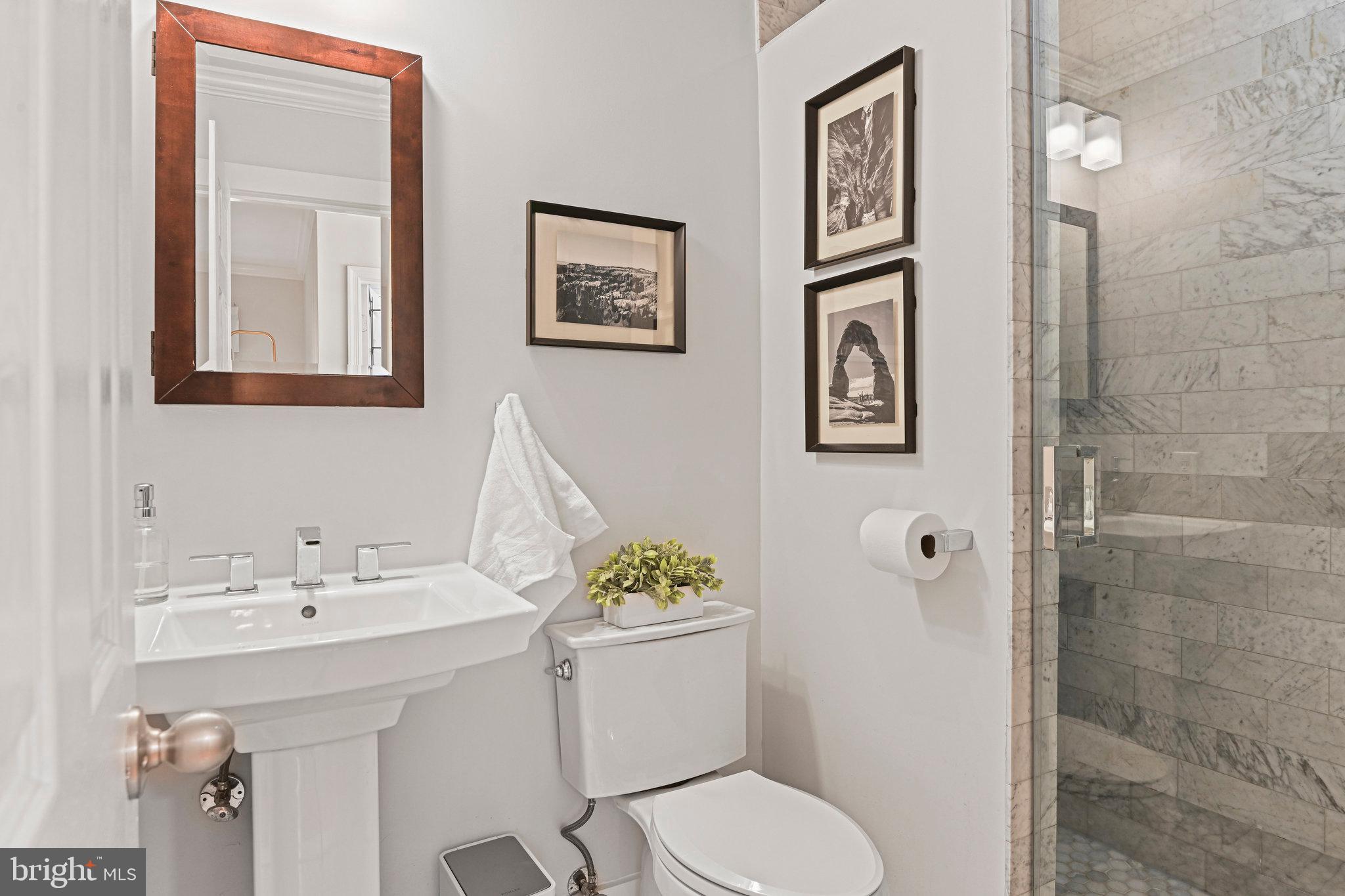 Bathroom with a marble-tiled glass shower, pedestal sink, and framed photographs.