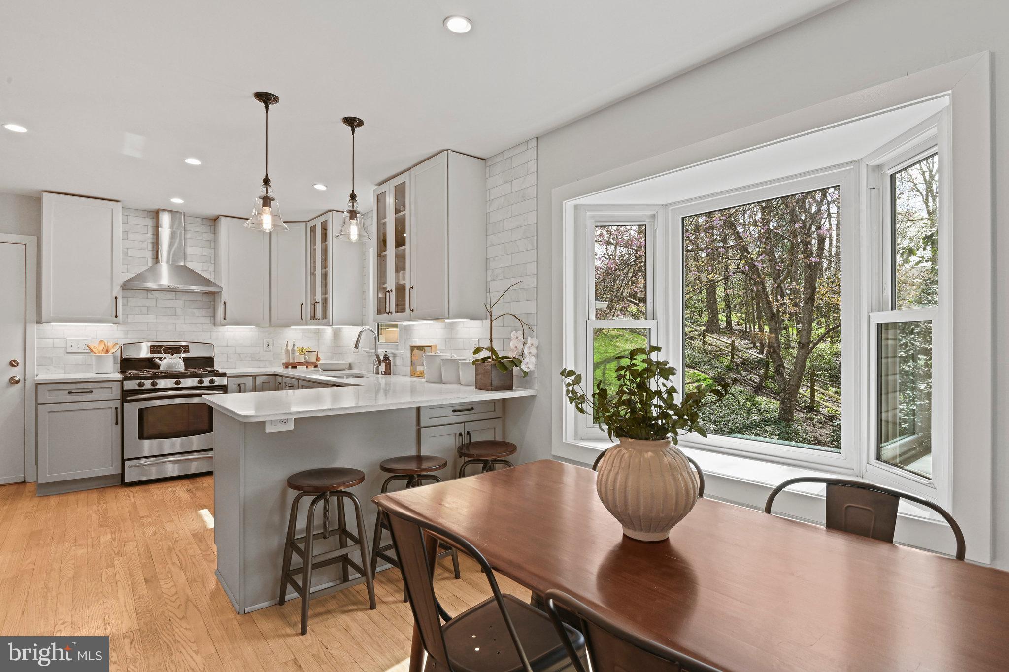 Bright kitchen and dining area with a white island, stainless steel range, and large bay window overlooking trees.