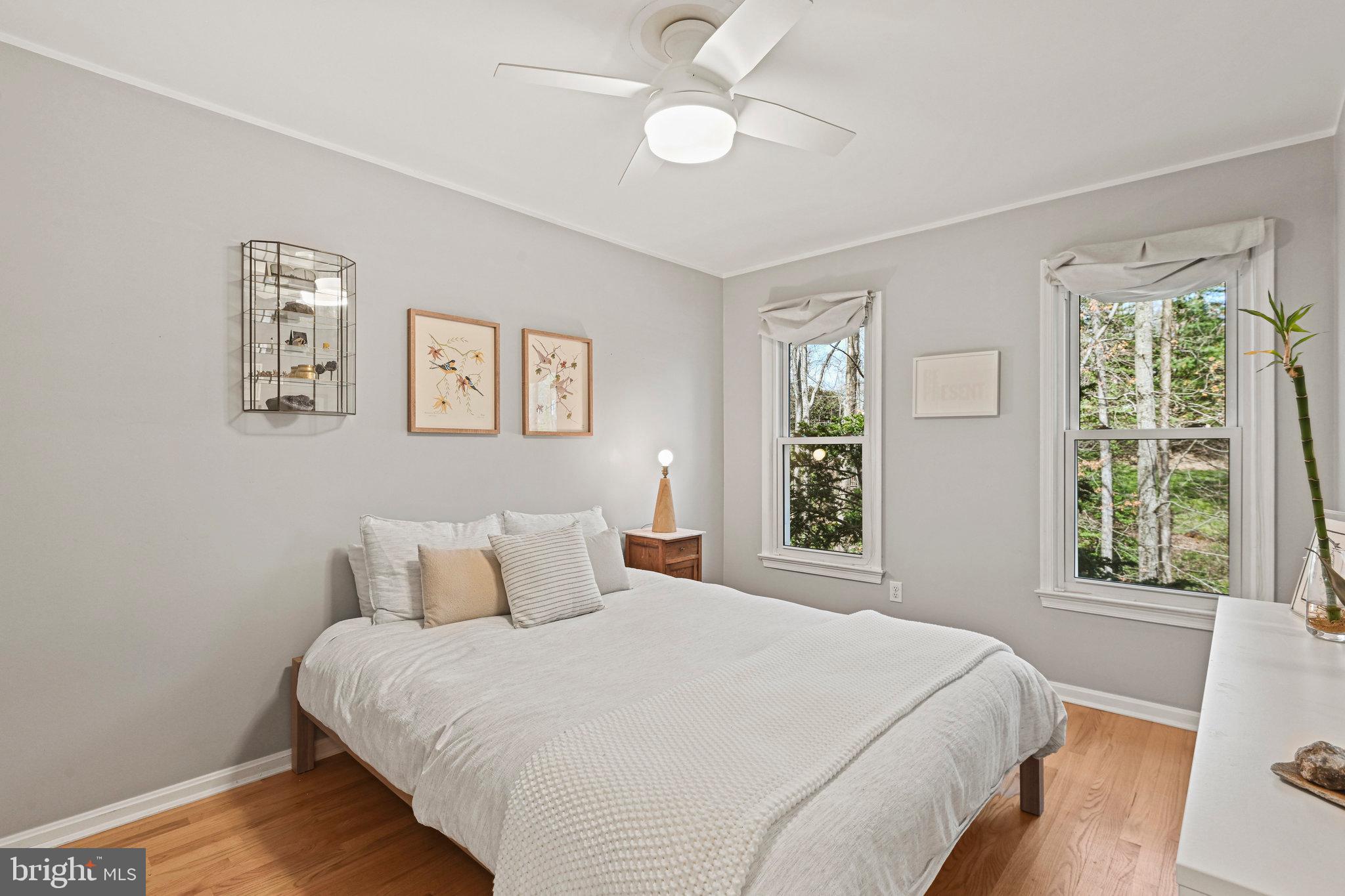 Staged bedroom with a white bed, hardwood floors, and windows overlooking trees.