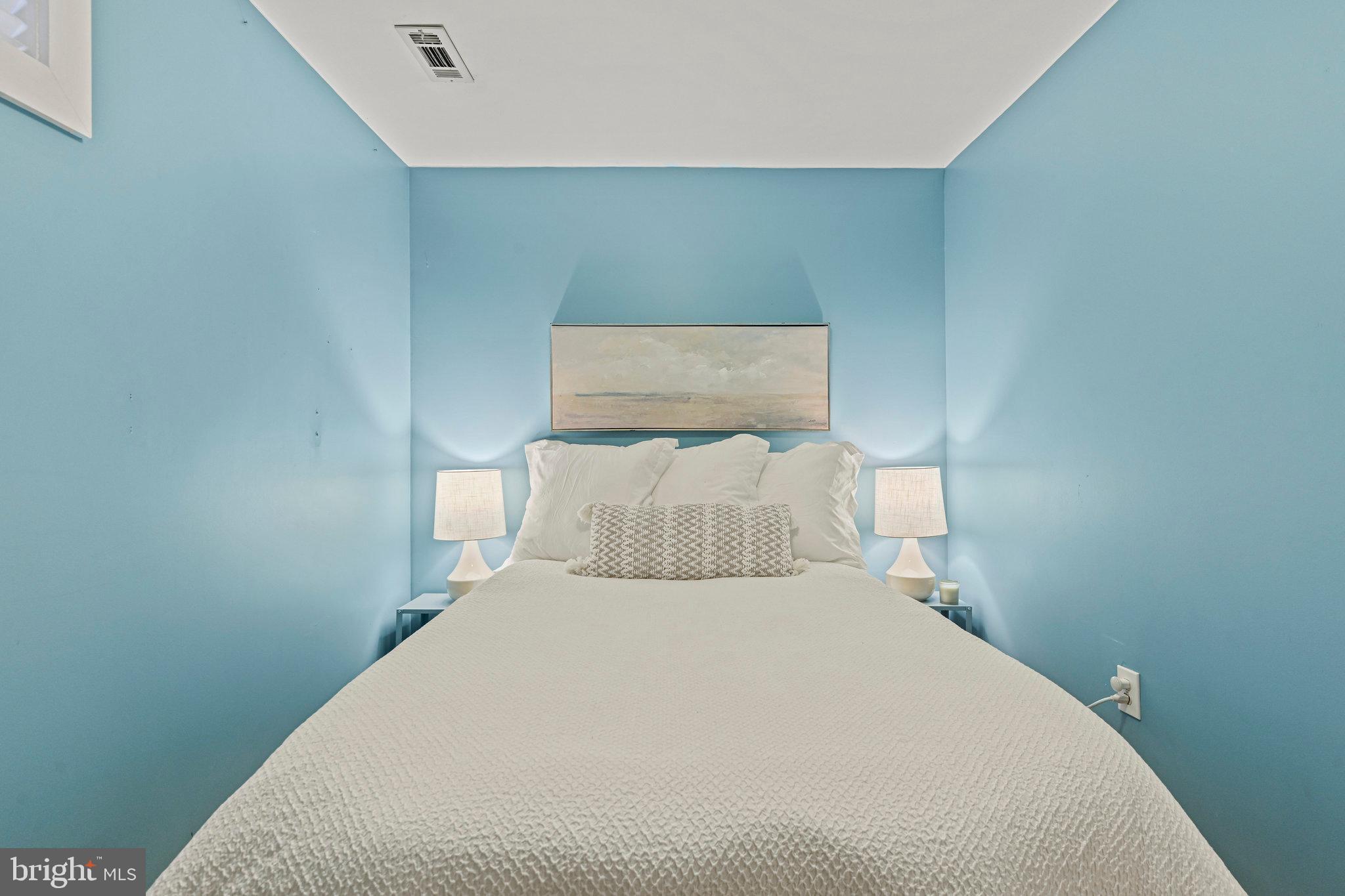 Staged bedroom with a white-covered bed, blue walls, matching bedside lamps, and a landscape print.
