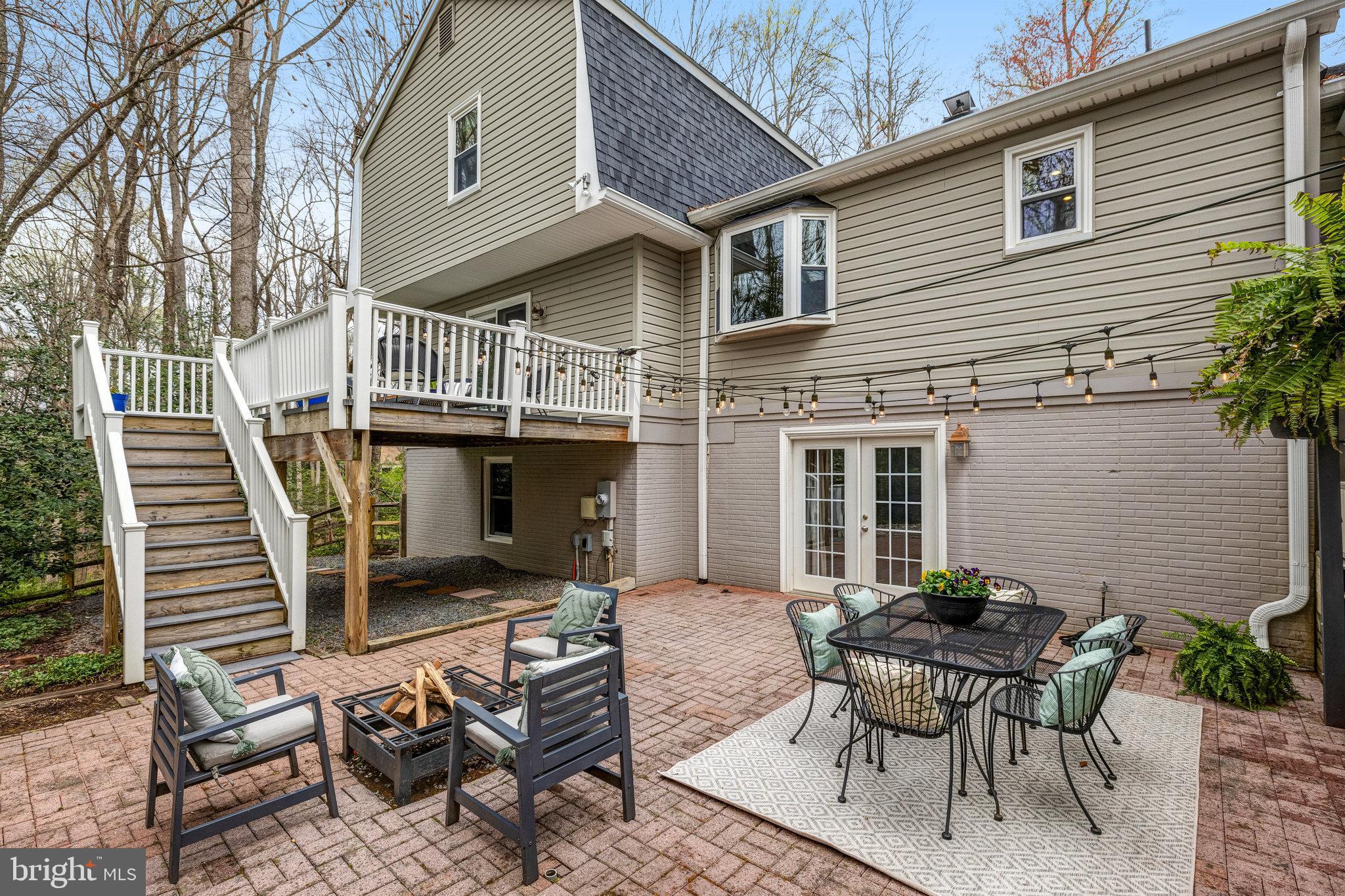 Brick patio with outdoor dining and lounge seating, fire pit, string lights, and stairs to an elevated deck.