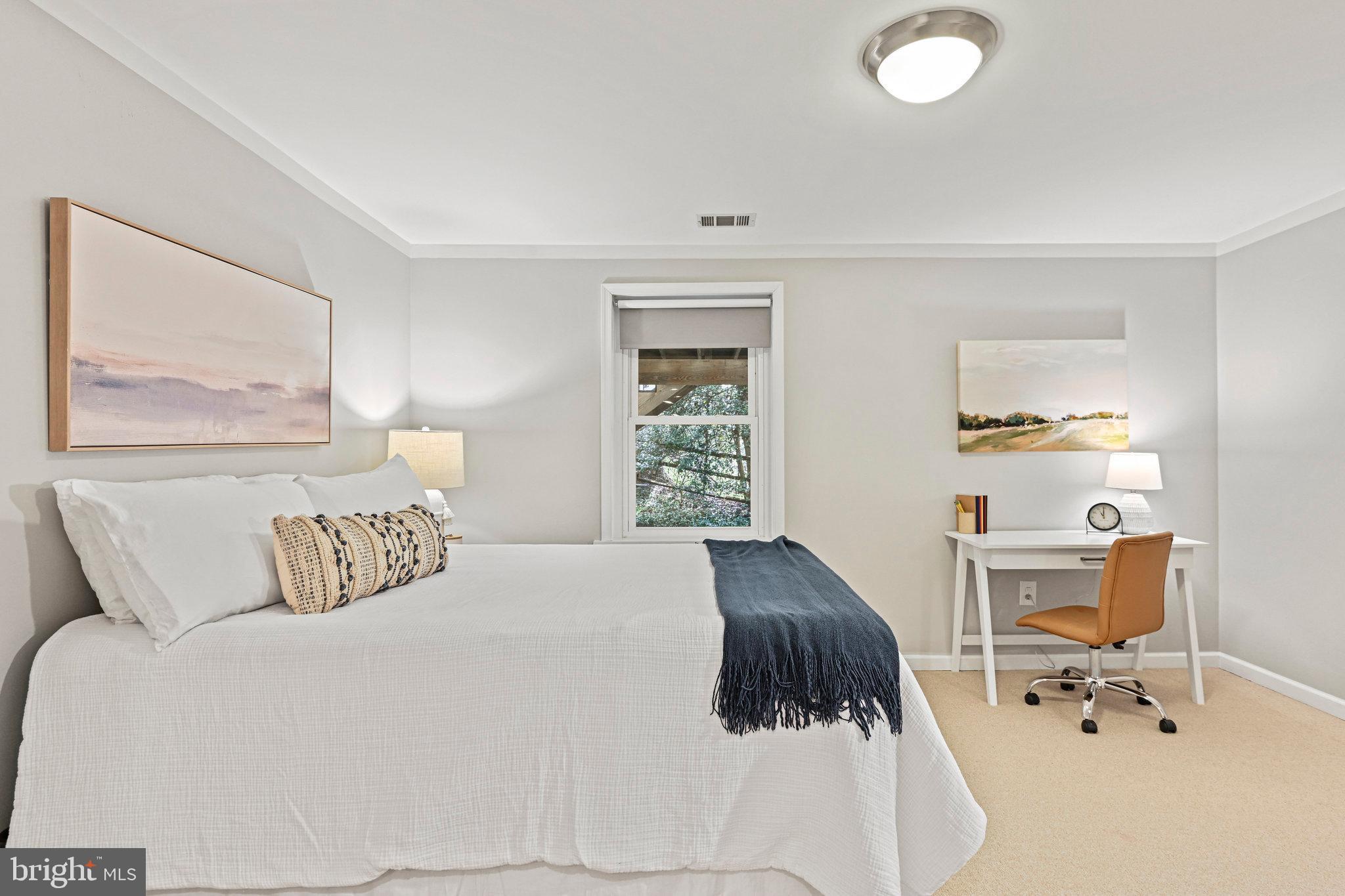 Staged bedroom with a large white bed, navy throw, and desk beside a window.