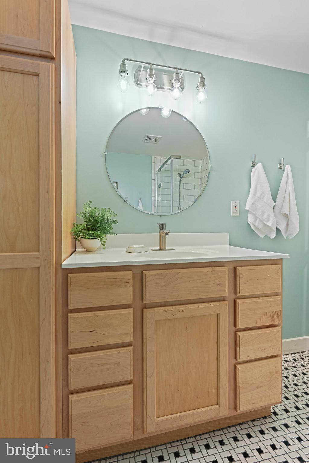 Bathroom vanity with natural wood cabinetry, pale aqua walls, a round mirror, and black-and-white mosaic flooring.