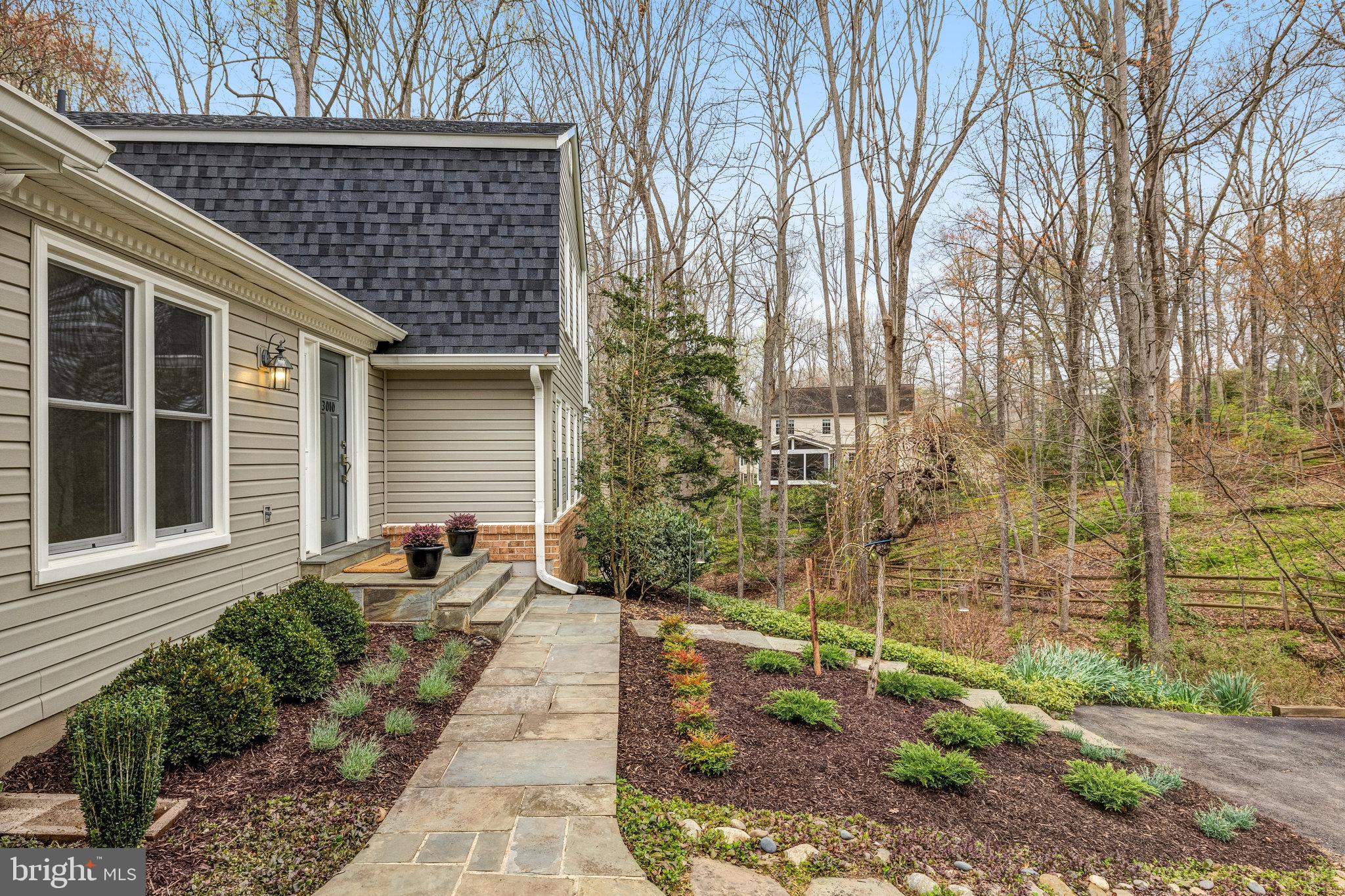 Home entrance with stone walkway, landscaped garden beds, and wooded yard.