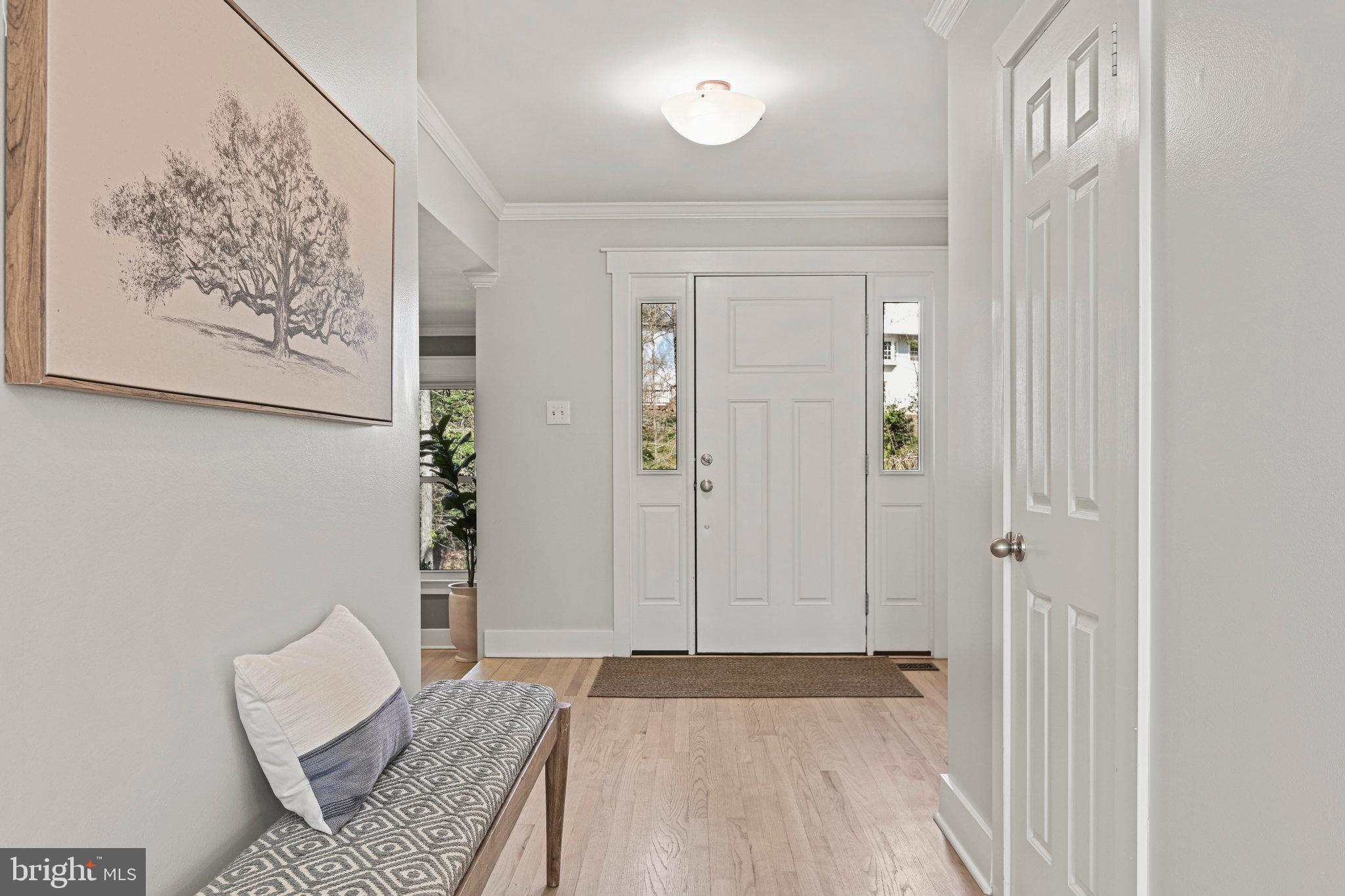 Staged entryway with a white front door, narrow sidelights, light hardwood floors, and a patterned bench.