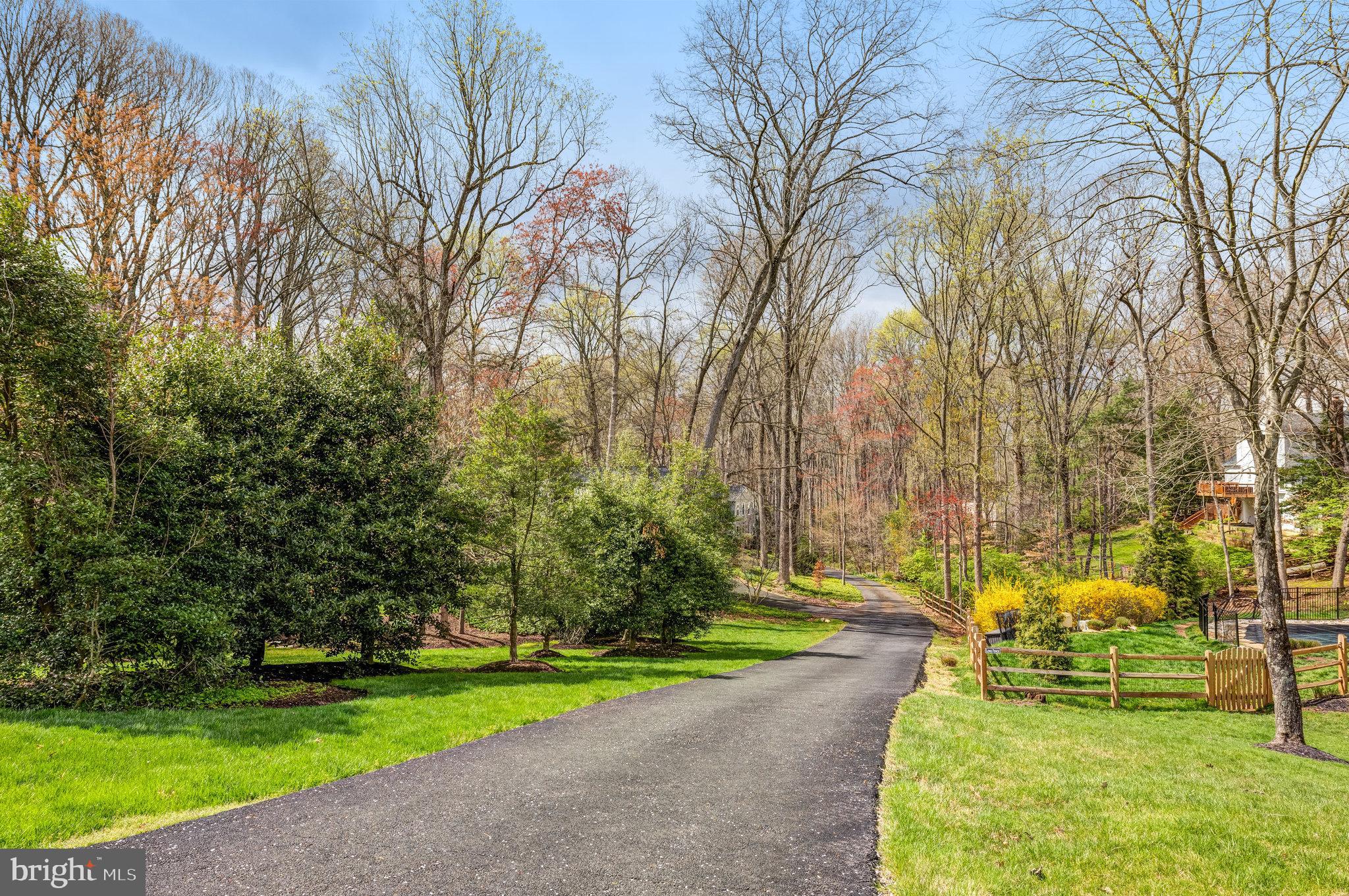 Curving paved driveway through wooded grounds, bordered by green lawns, shrubs, and a wooden fence.