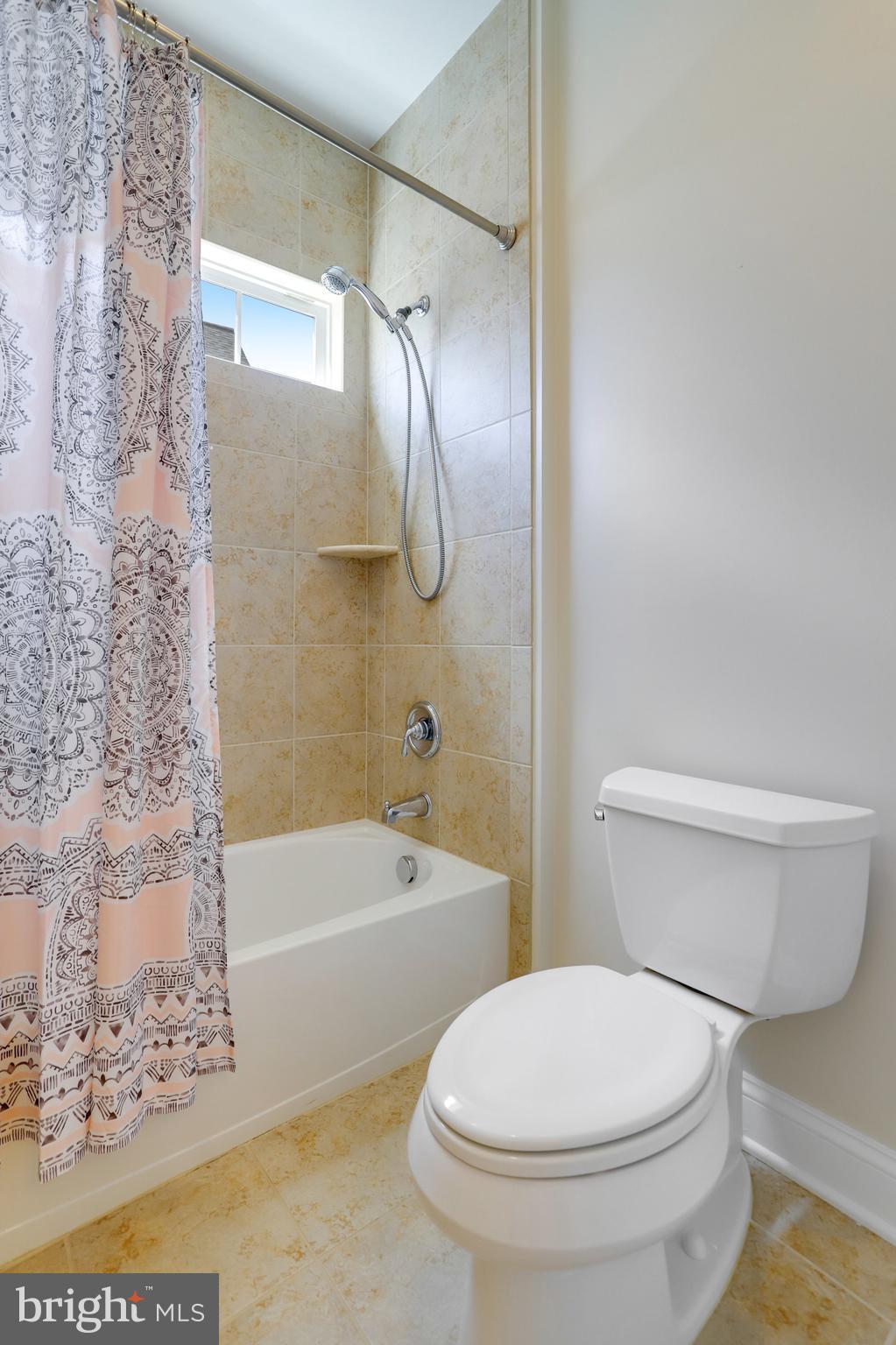 Bathroom with a tiled tub and handheld shower, small window, and patterned curtain.