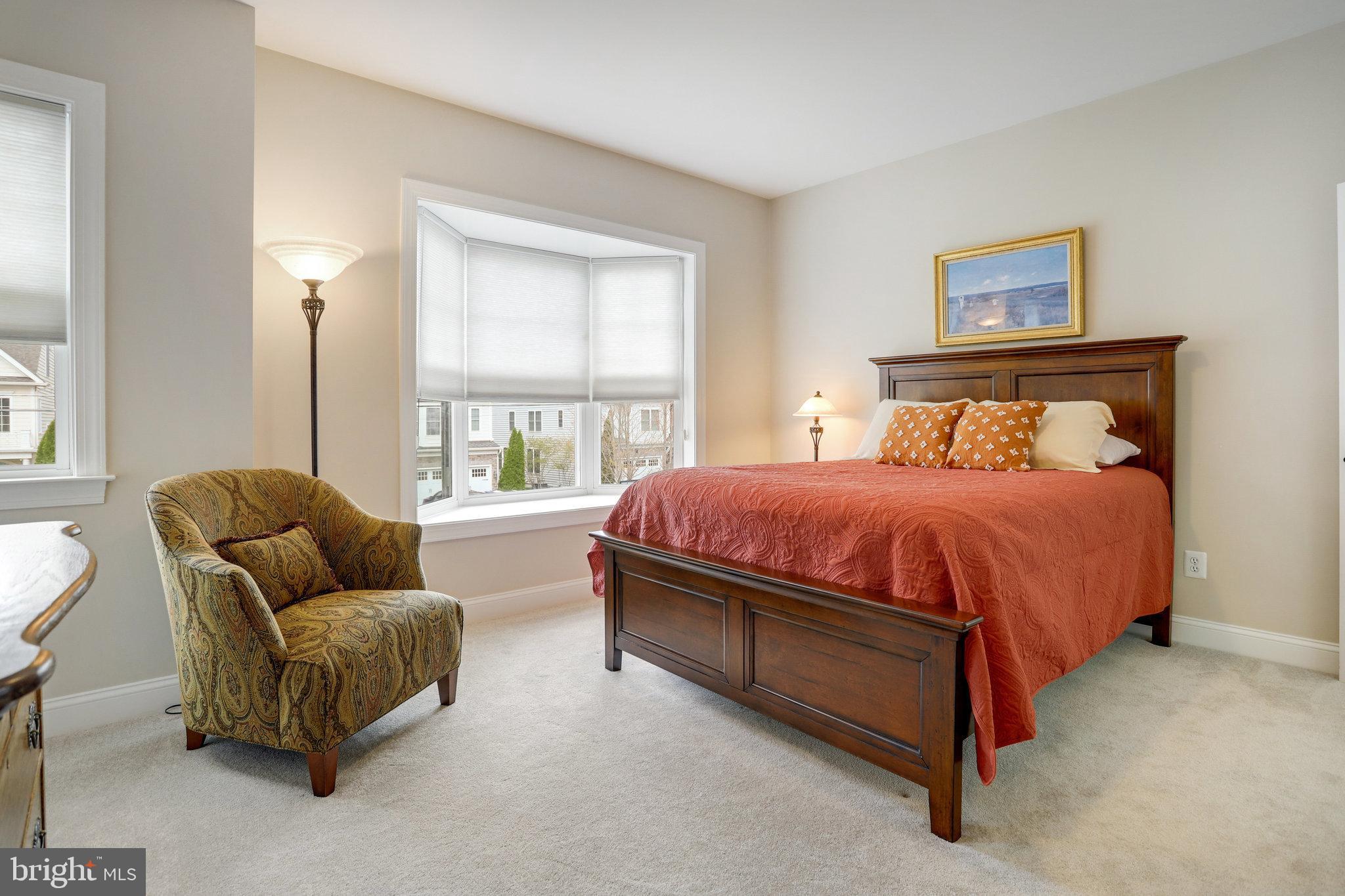 Furnished bedroom with a dark wood bed, rust-red quilt, patterned armchair, and wide windows.