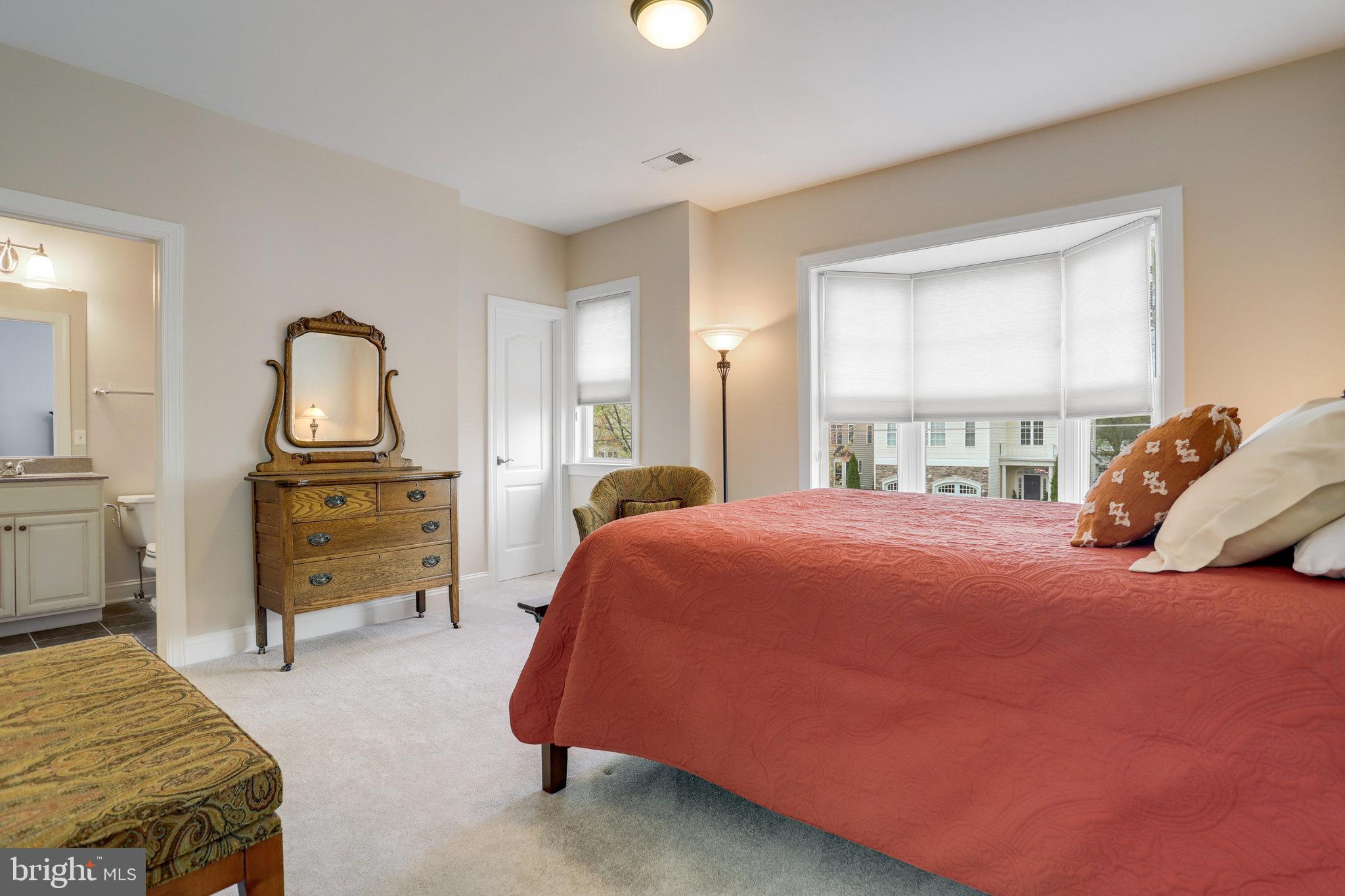 Bedroom with coral quilted bed, bay window, antique dresser and mirror, and an attached bathroom.