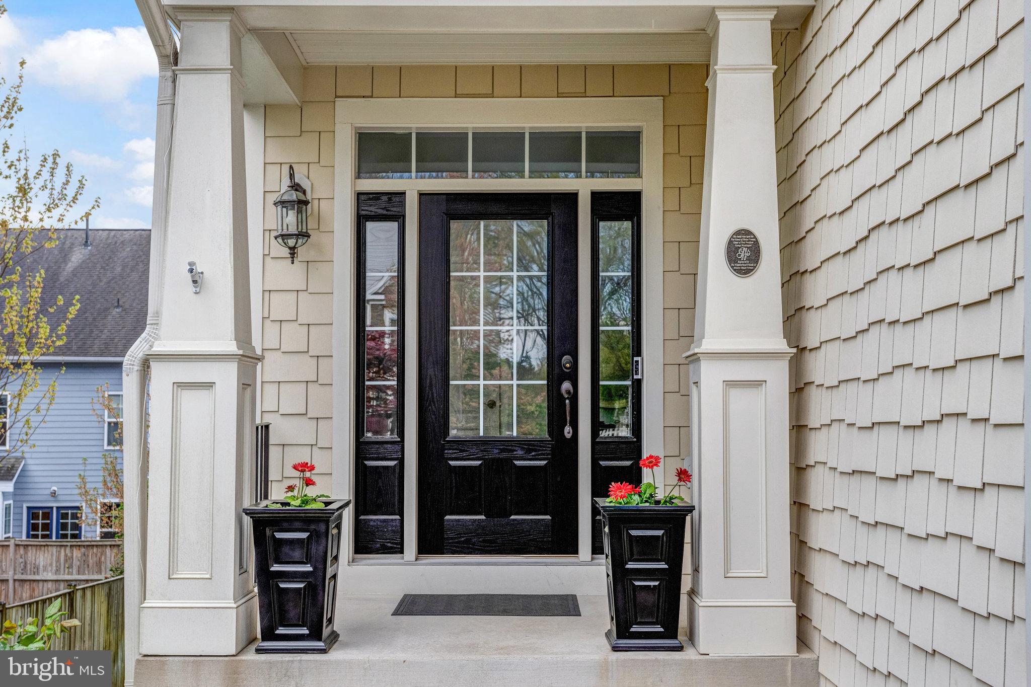 Covered front entrance with a black glass-paneled door, white columns, and red flowers in planters.