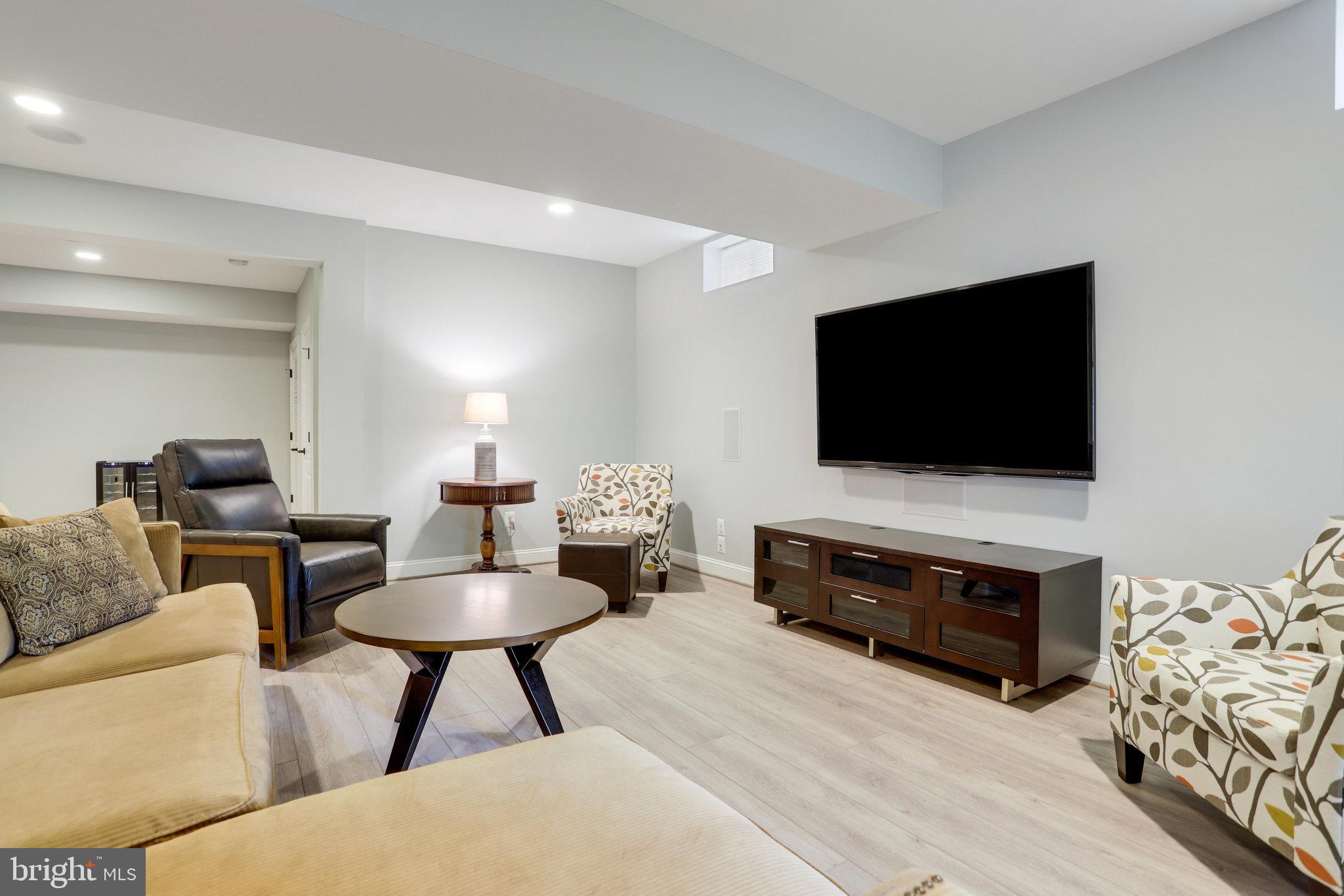 Furnished living room with a large wall-mounted TV, beige sofa, patterned armchairs, and pale wood flooring.