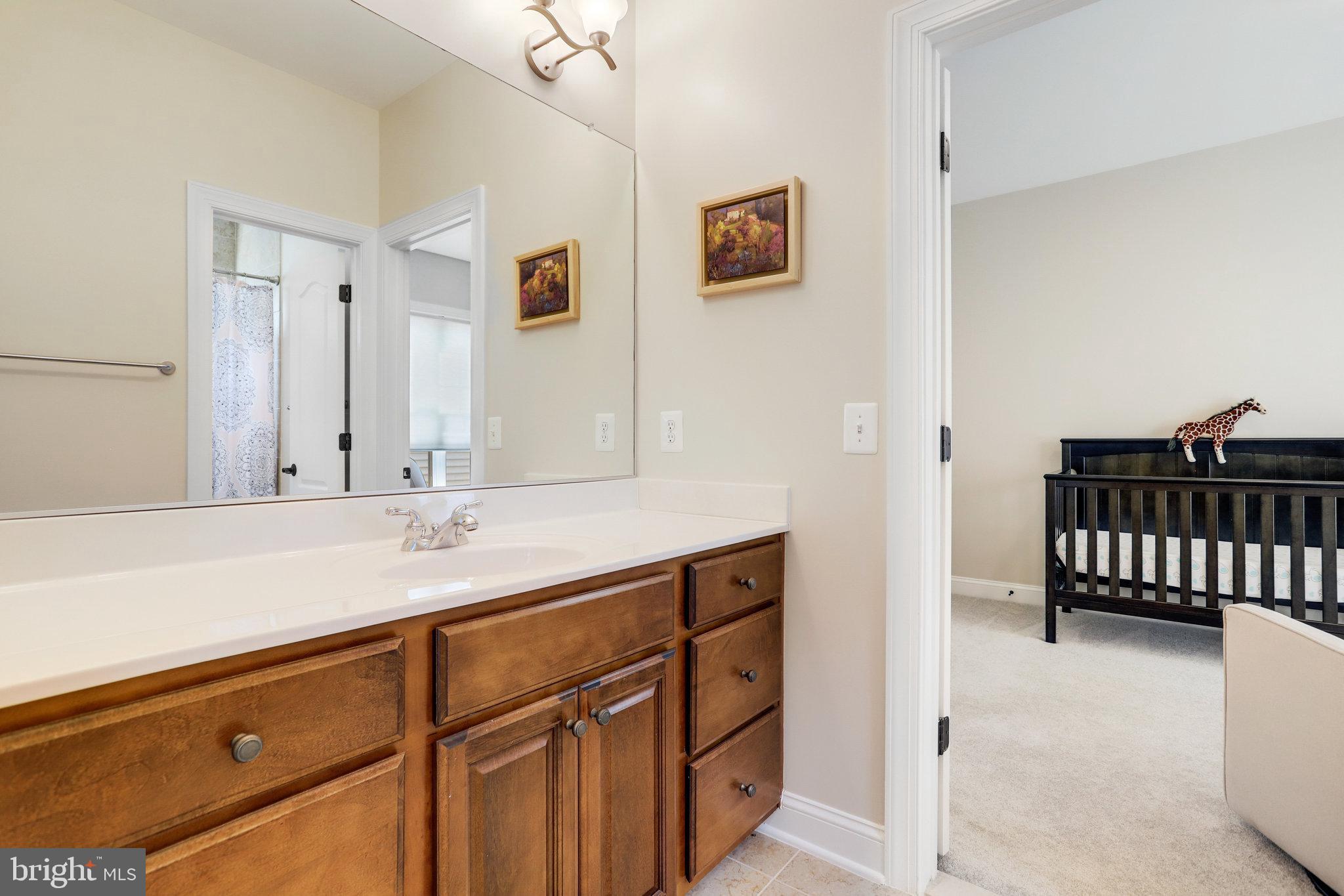 Bathroom with a wood vanity and large mirror, looking into a nursery with a dark crib.