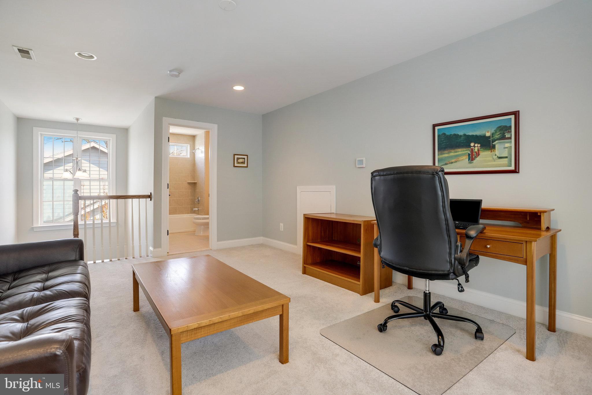 Furnished home office with a wooden desk, black chair, leather sofa, and doorway to a bathroom.