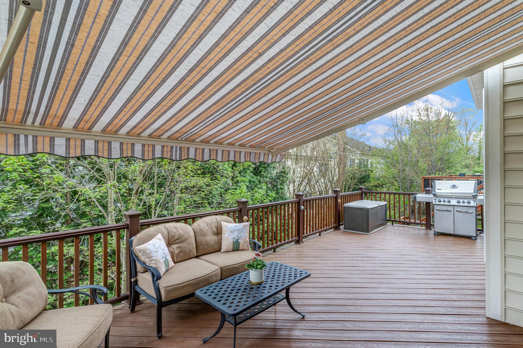Covered wooden deck with cushioned seating, a barbecue grill, and wooded surroundings.