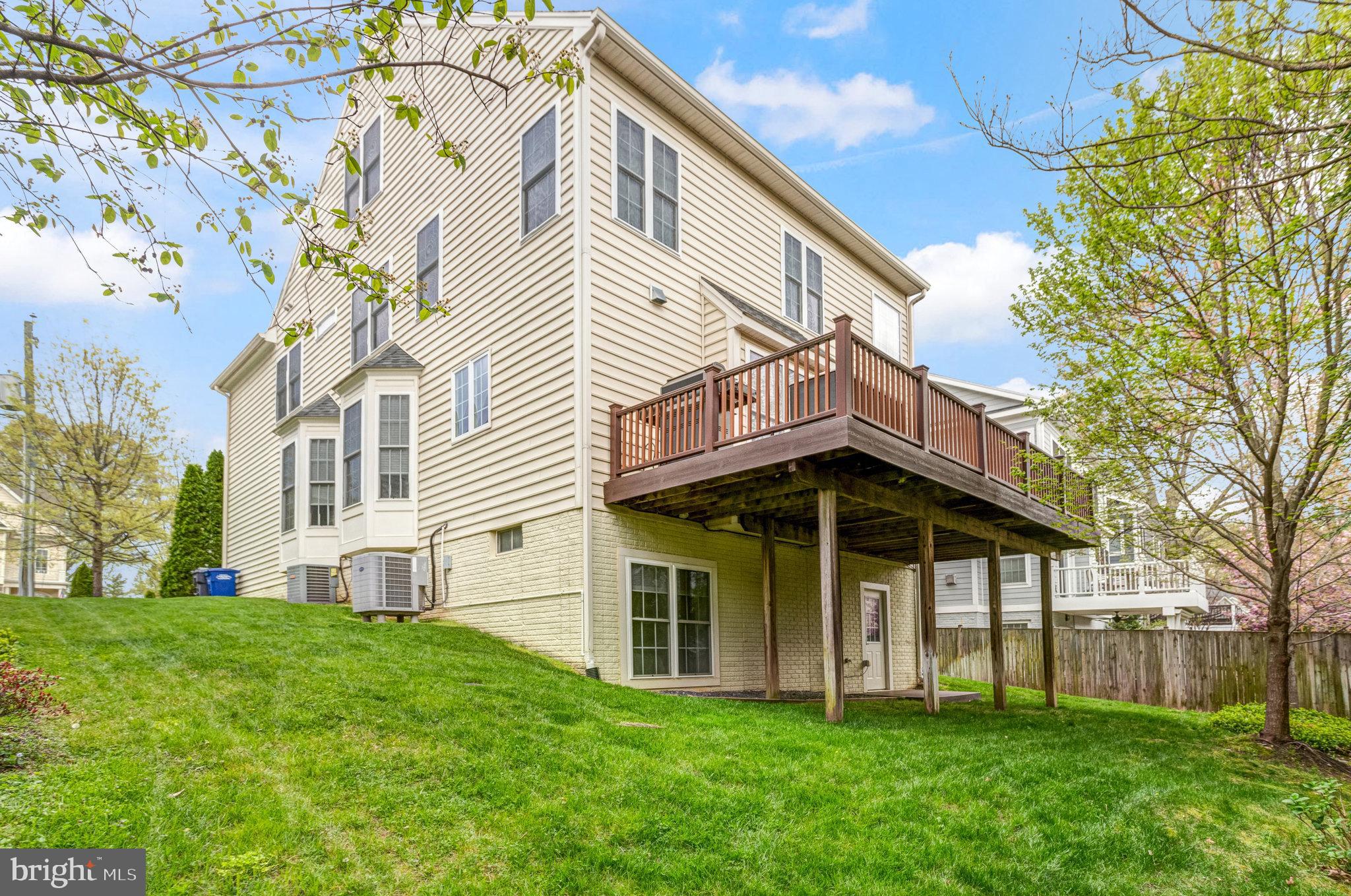 Rear of a beige three-story house with a raised wooden deck overlooking a sloping lawn.