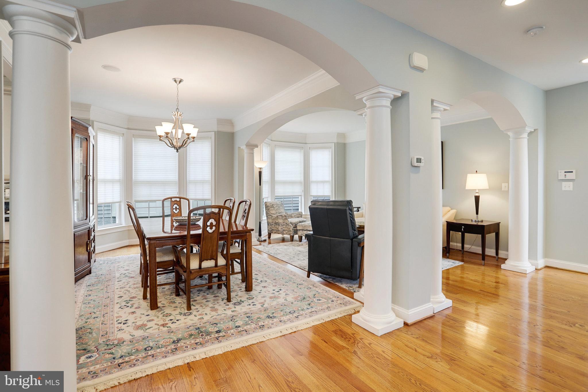 Dining and living area with hardwood floors, white columns, arched openings, and a wooden dining set by bay windows.