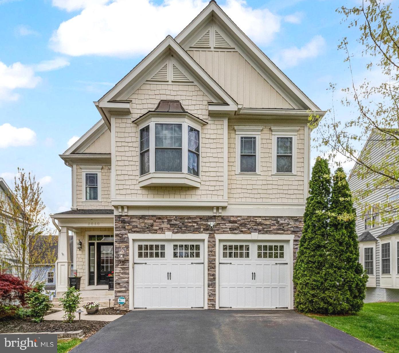 Two-story house with steep gables, stone facade, twin white garage doors, and a tall evergreen by the driveway.