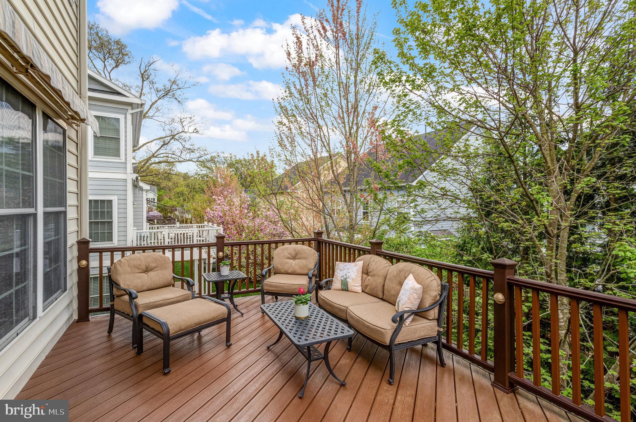 Furnished wooden deck with cushioned seating overlooking leafy trees and neighboring homes.