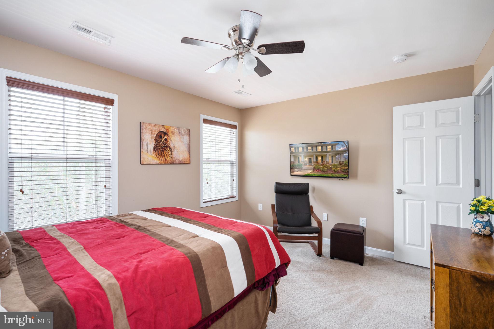Furnished bedroom with a striped red-and-brown bedspread, two windows, and a black chair beneath a wall-mounted television.