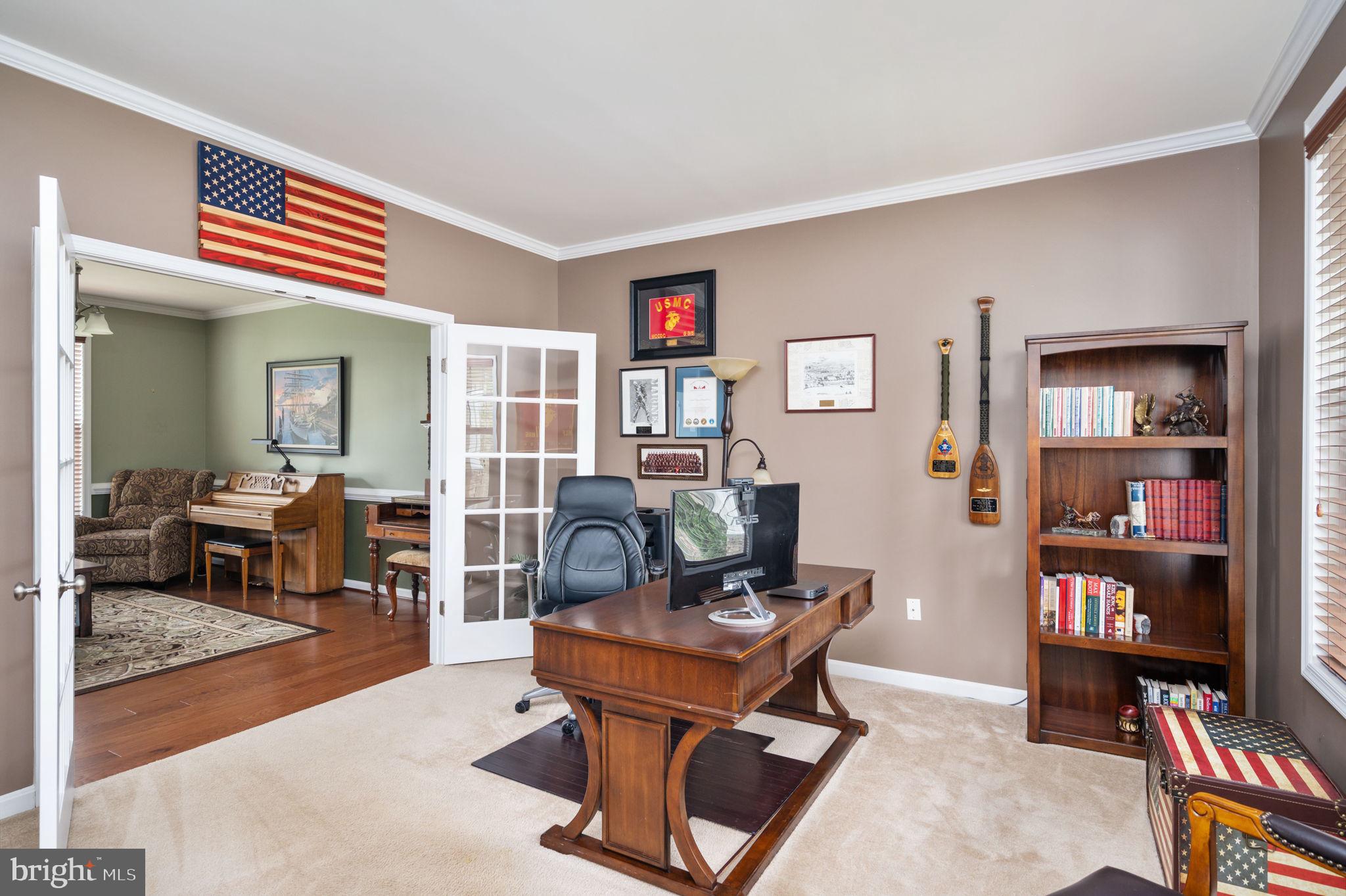 Home office with a wooden desk, bookcase, and French doors opening to a sitting room with a piano.
