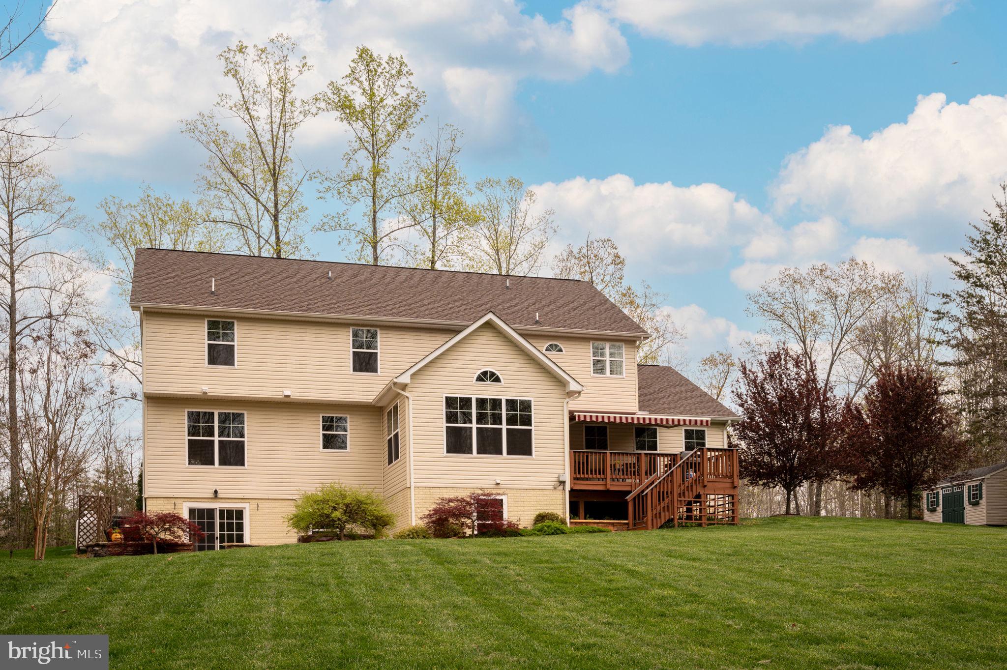 Rear of a two-story beige home with a raised wooden deck, striped awning, and broad lawn.