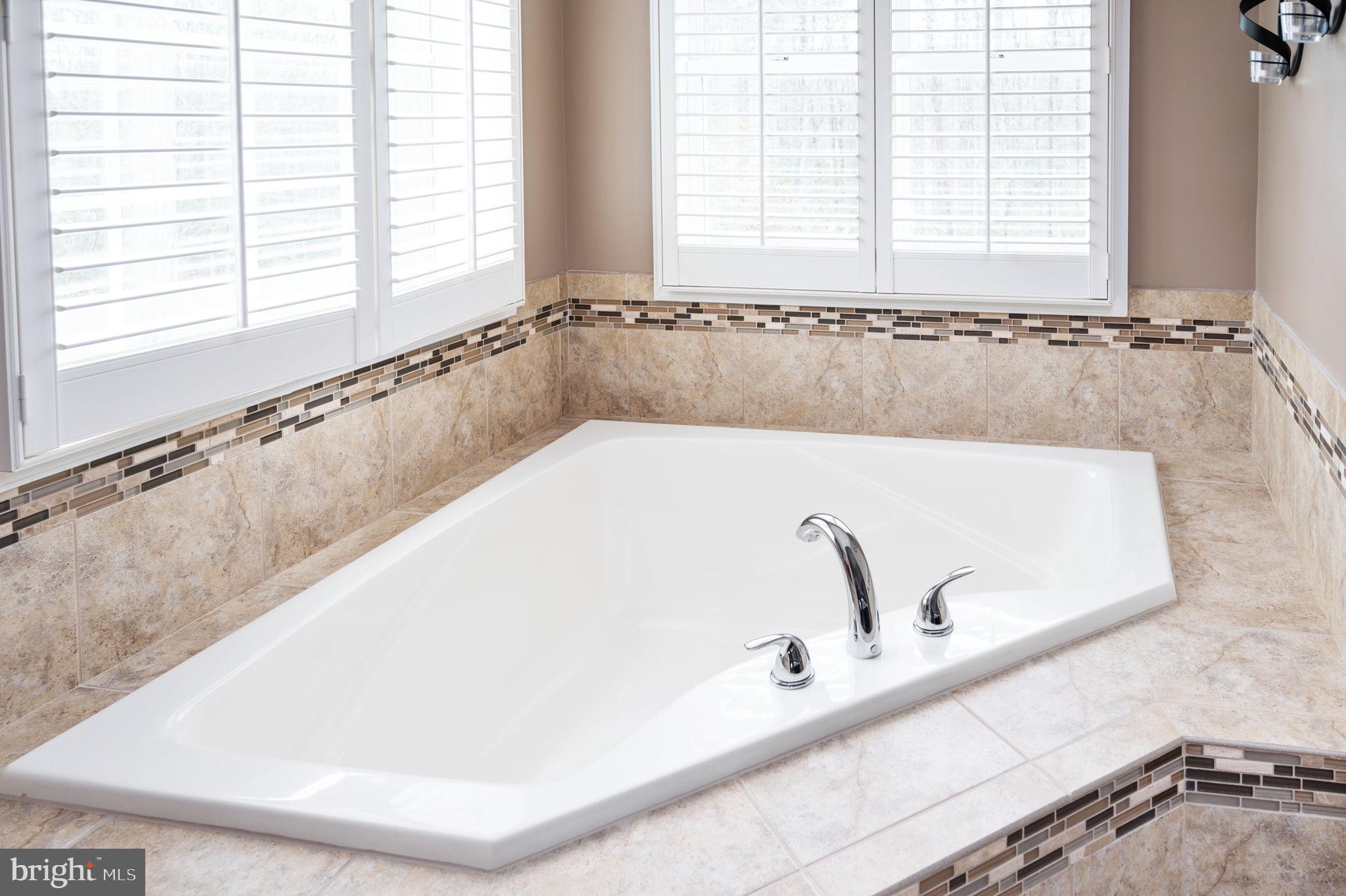 Bathroom with a large corner bathtub, chrome fixtures, tiled surround, and shuttered windows.