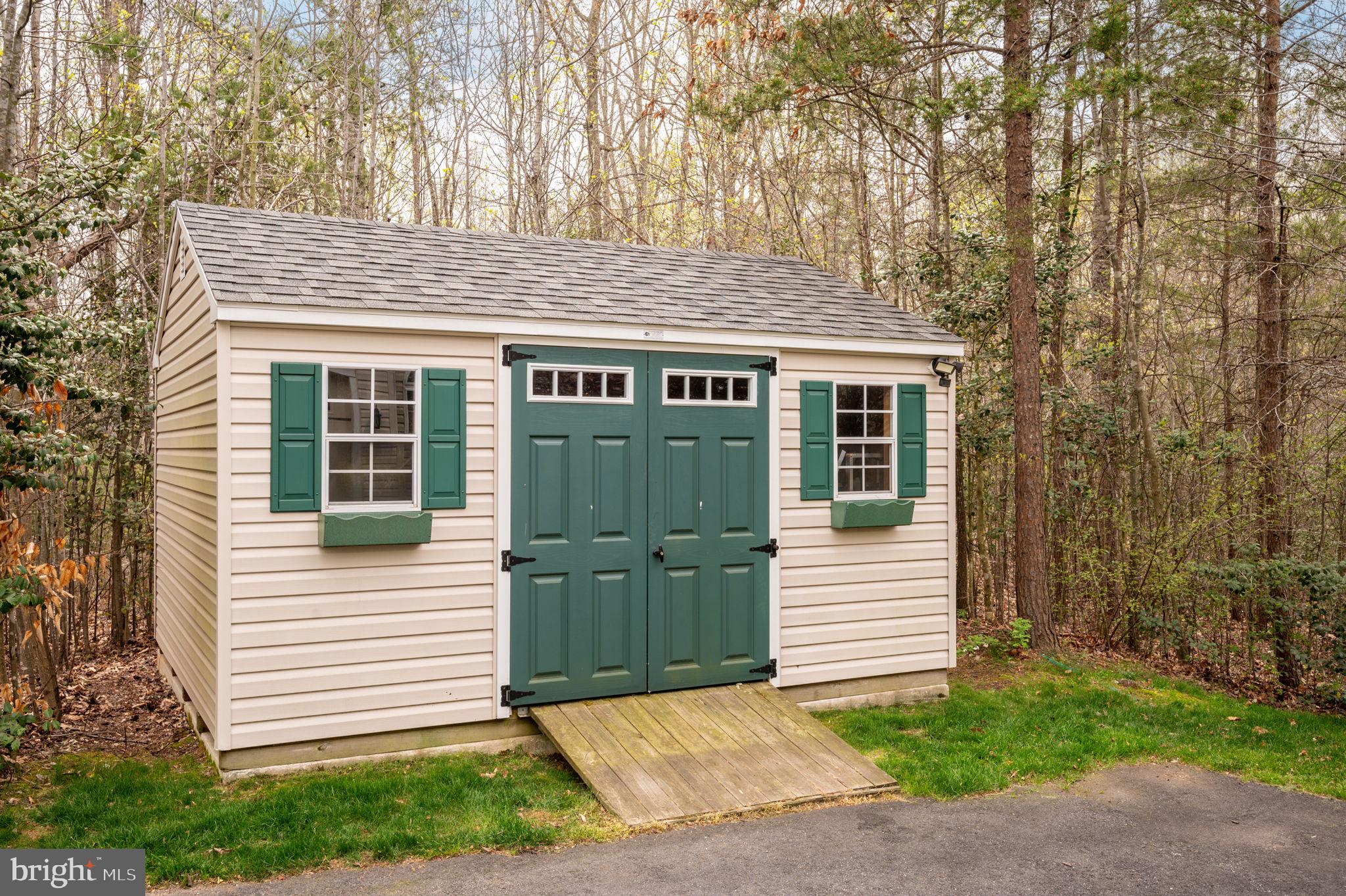 Cream storage shed with green double doors and shutters, set among trees beside a driveway.