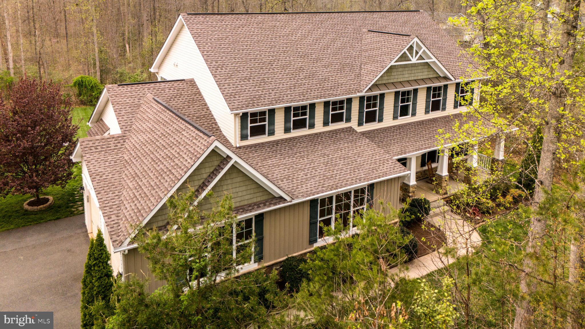 Aerial view of a beige two-story home with multiple gabled roofs, dark shutters, and a wooded yard.