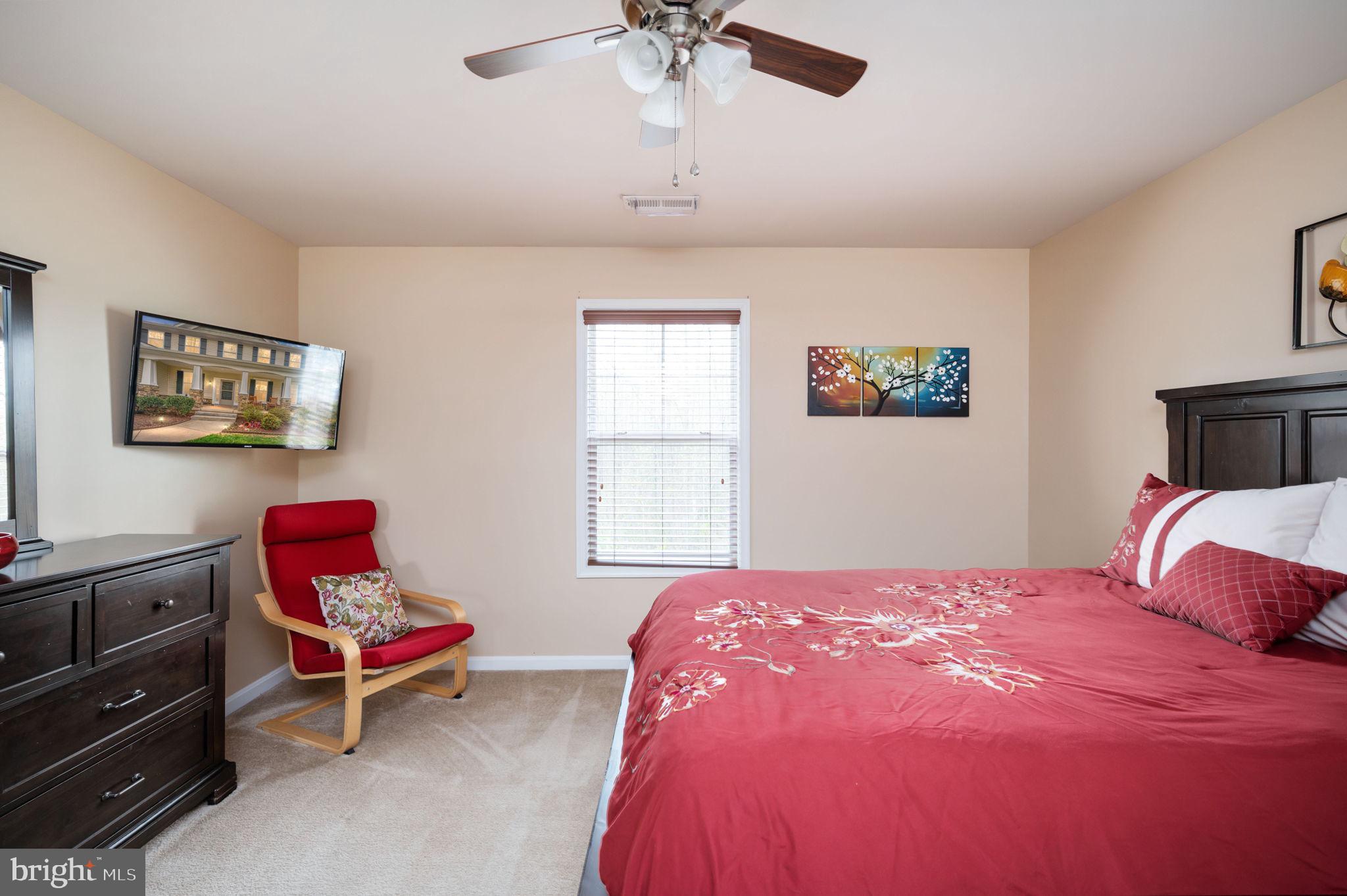 Bedroom with a red floral bedspread, red accent chair, dark wood dresser, and ceiling fan.