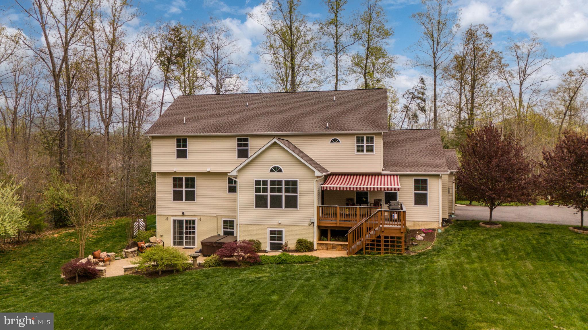 Rear of a two-story home with a raised deck, striped awning, landscaped lawn, and wooded surroundings.