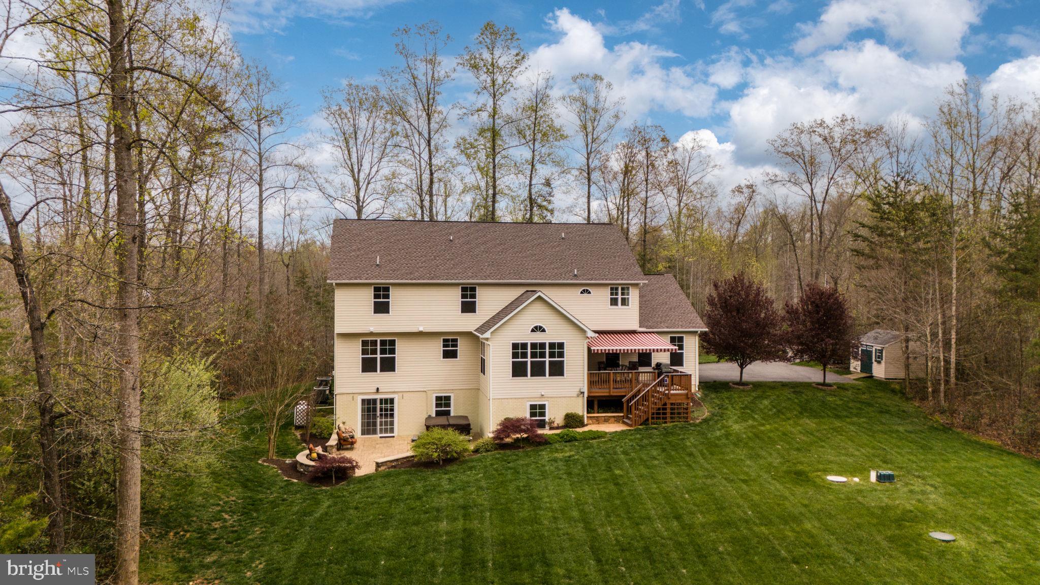 Aerial rear view of a beige two-story house with a deck, striped awning, patio, and expansive lawn.