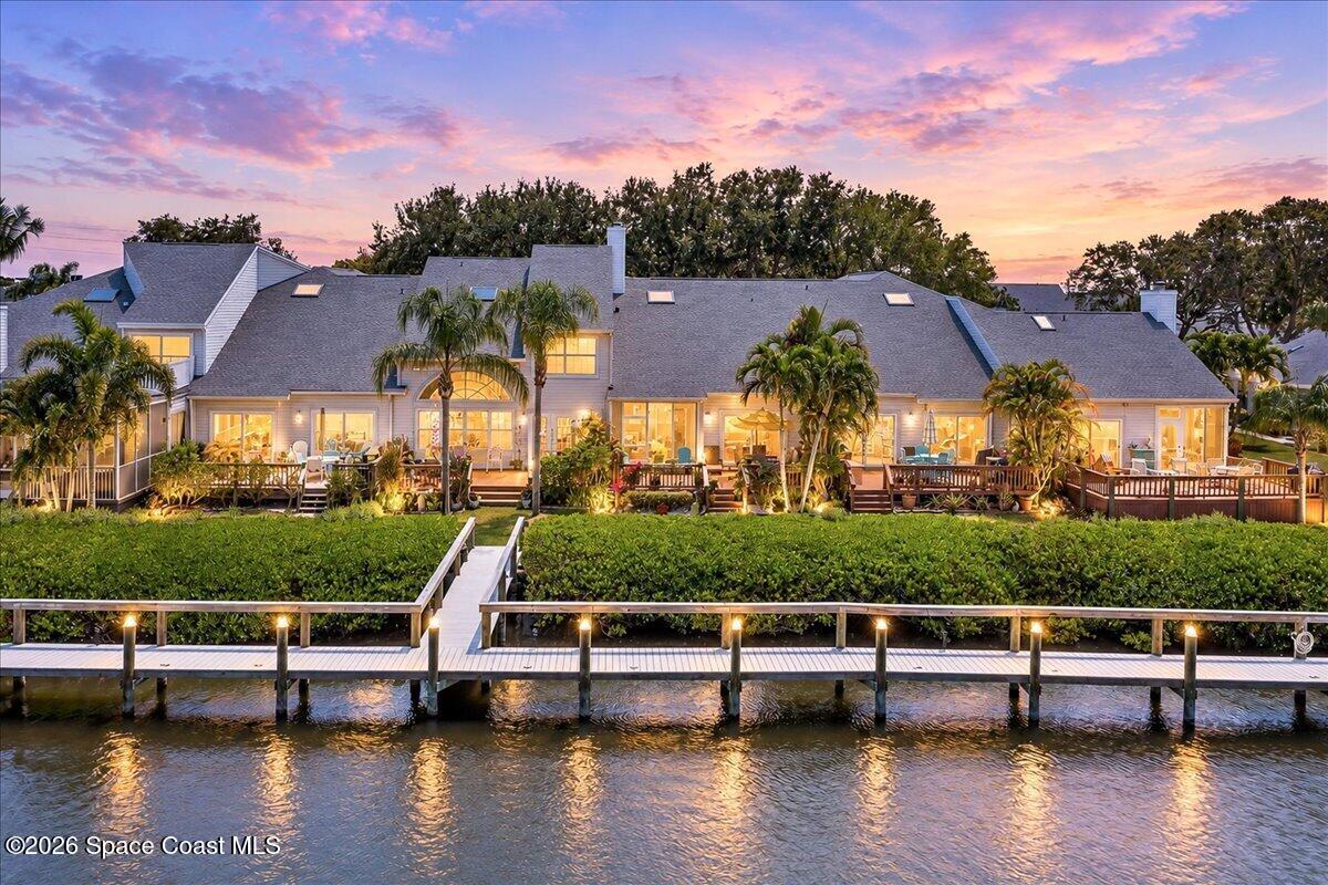 Waterfront home with a long dock, palm trees, and glowing windows beneath a pink dusk sky.