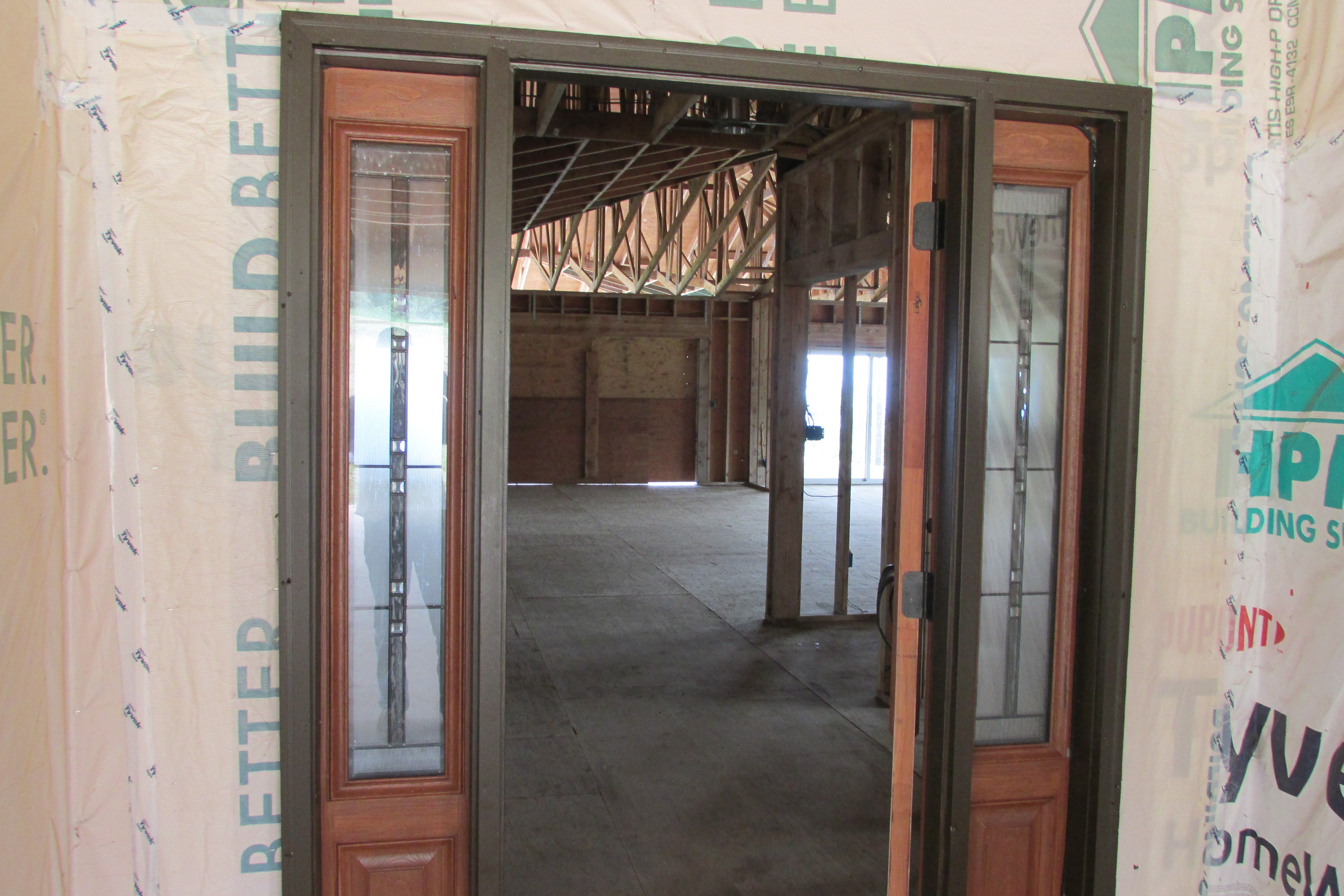 Unfinished home interior viewed through a glass-paneled entrance, with exposed roof trusses and framing.