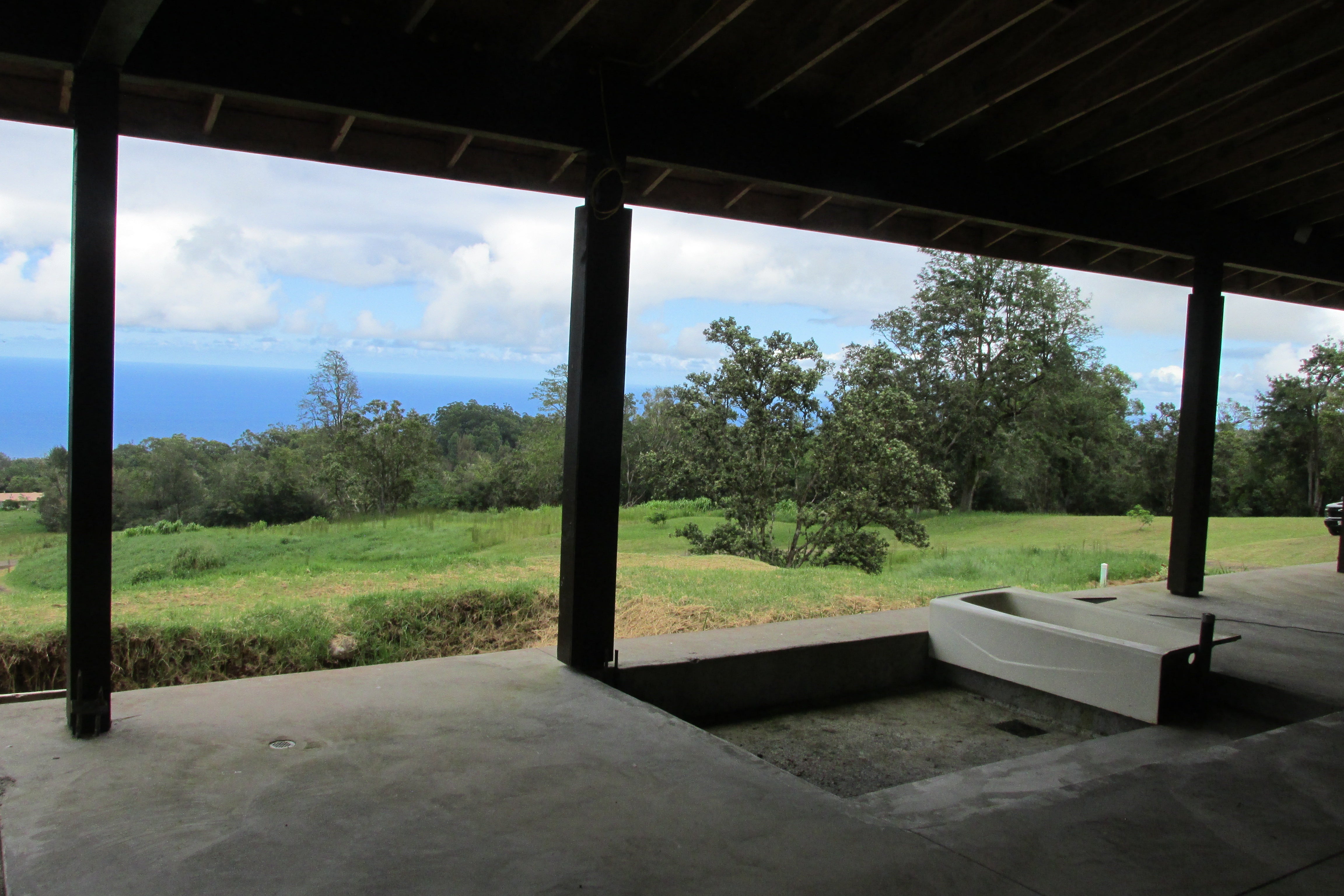 Covered concrete patio with a utility sink overlooking grassy grounds, trees, and the ocean.