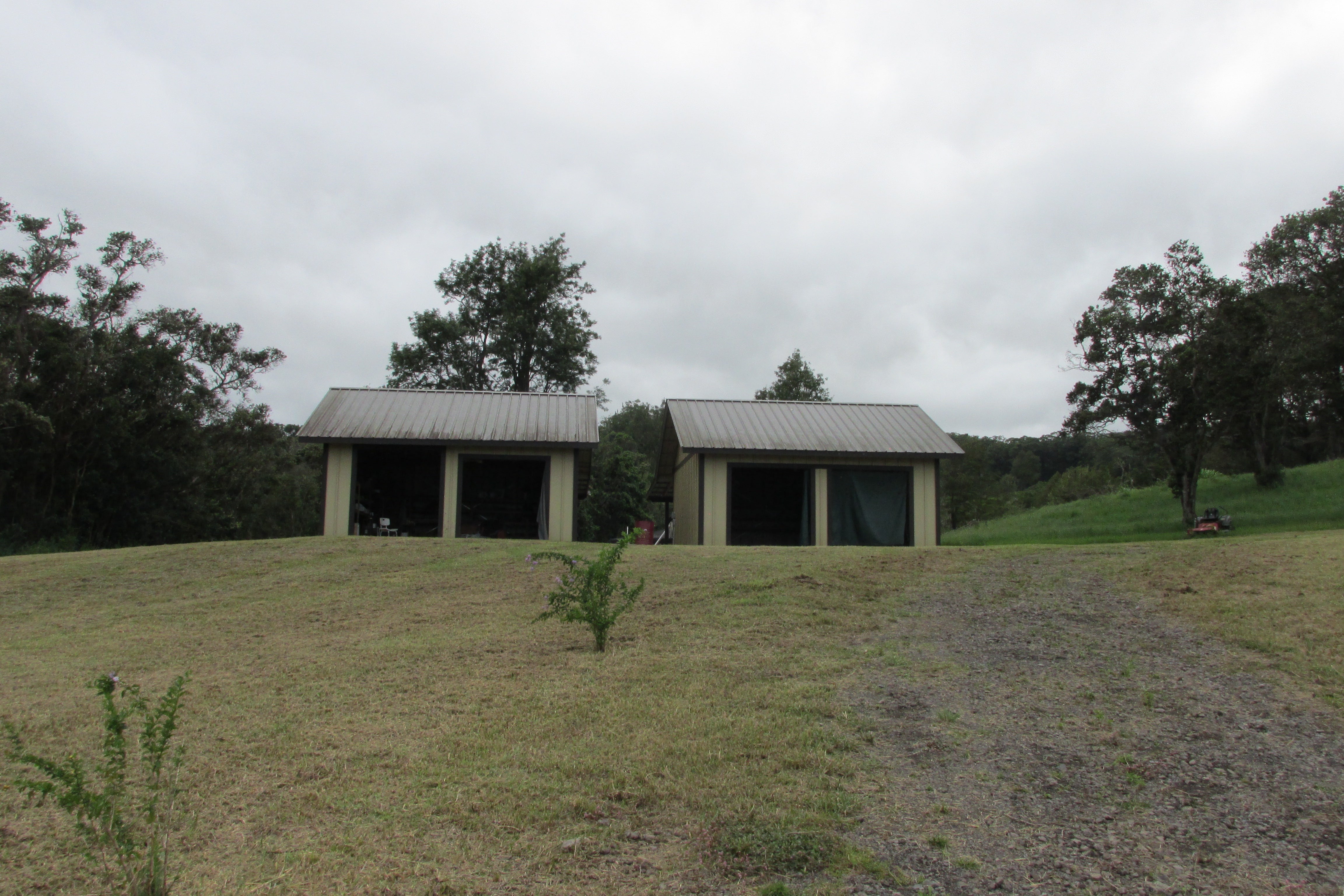 Two metal-roofed, open-front utility buildings on a grassy slope surrounded by trees.