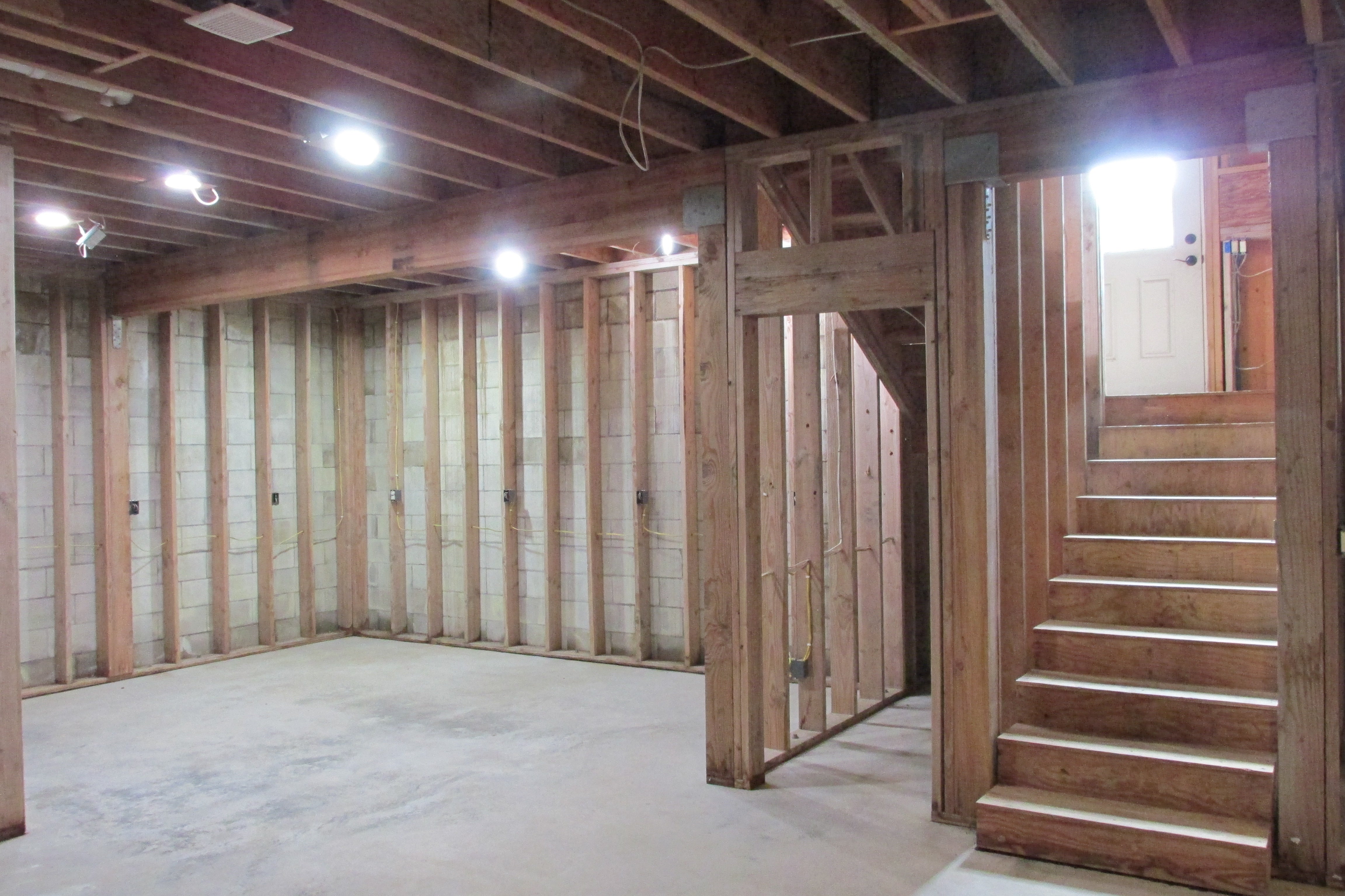 Unfinished basement with exposed framing, concrete floor, and wooden stairs leading to an upper door.