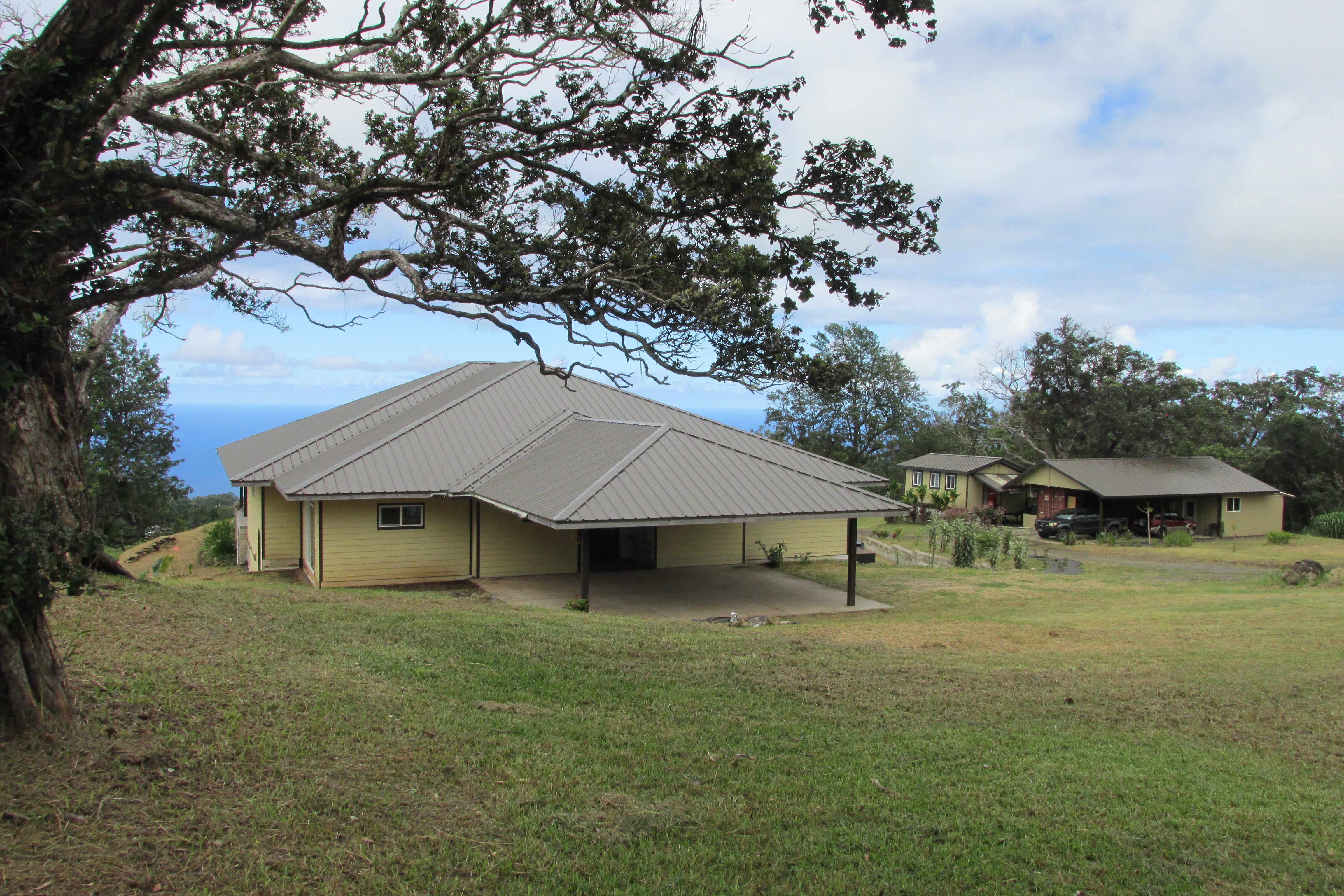 Tan home with a broad metal roof and covered carport, framed by trees with the ocean beyond.
