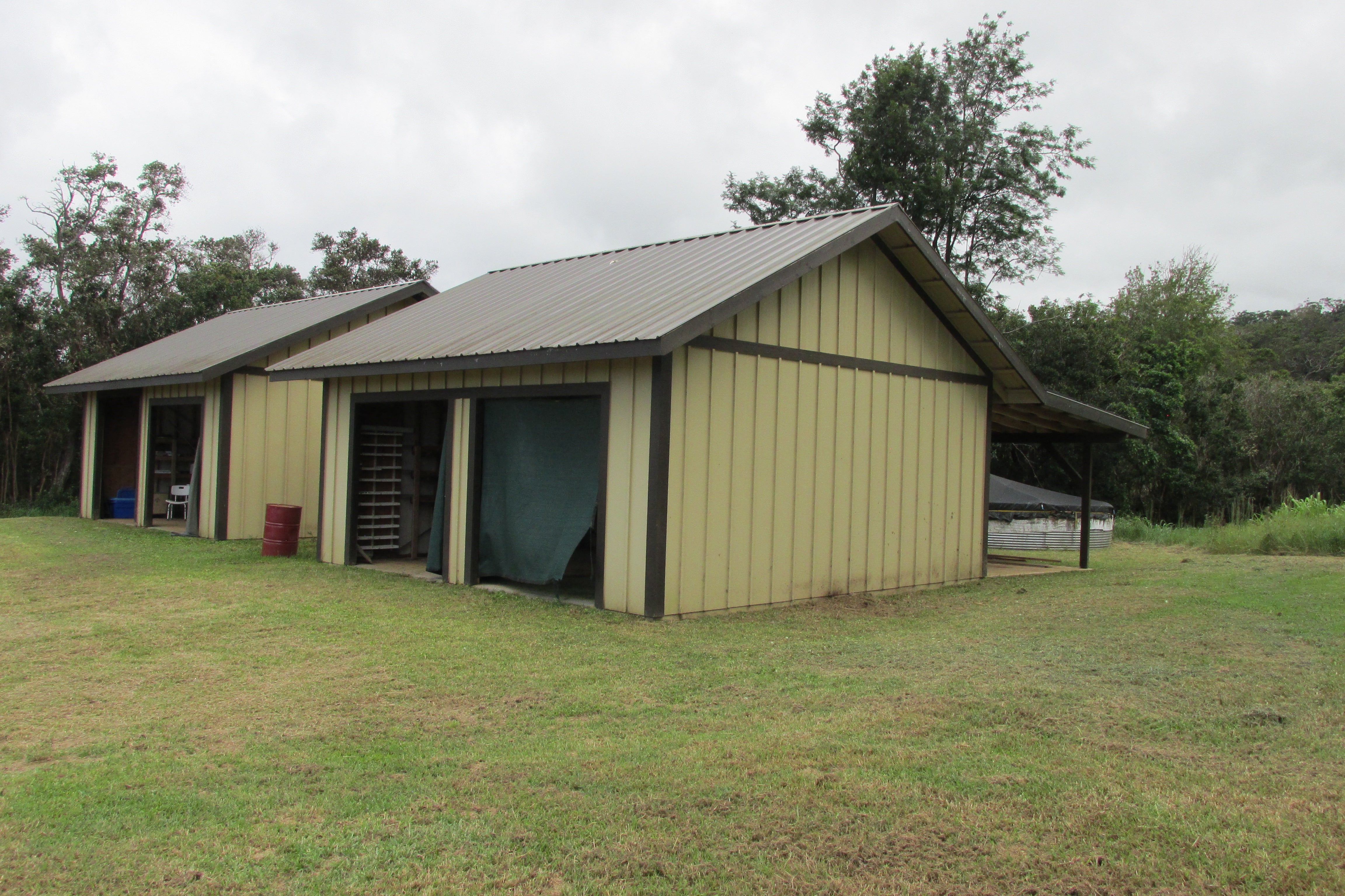 Tan agricultural outbuilding with open bays, a metal roof, and a round water tank beside it.