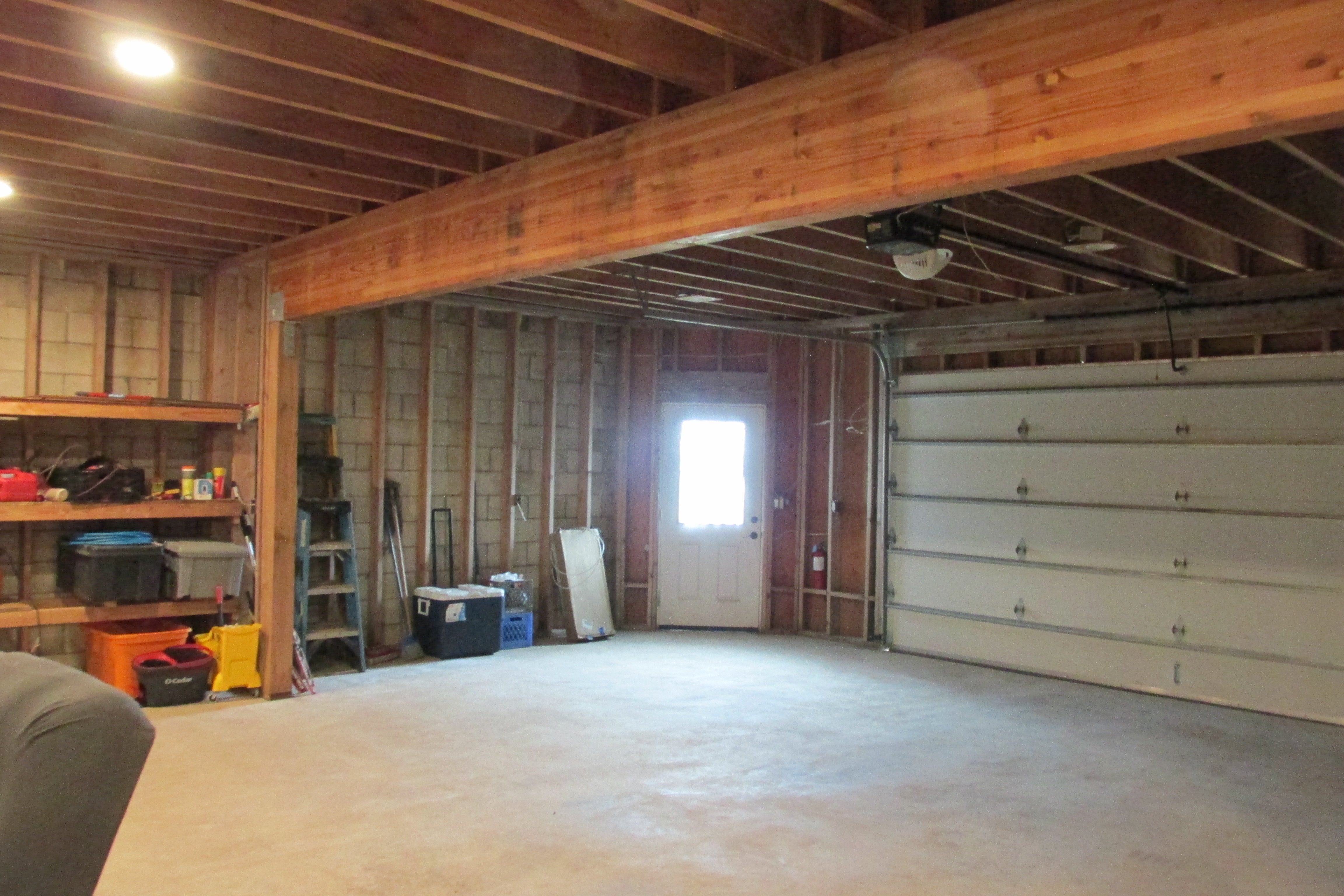 Garage with exposed framing, concrete floor, overhead door, and storage shelves.