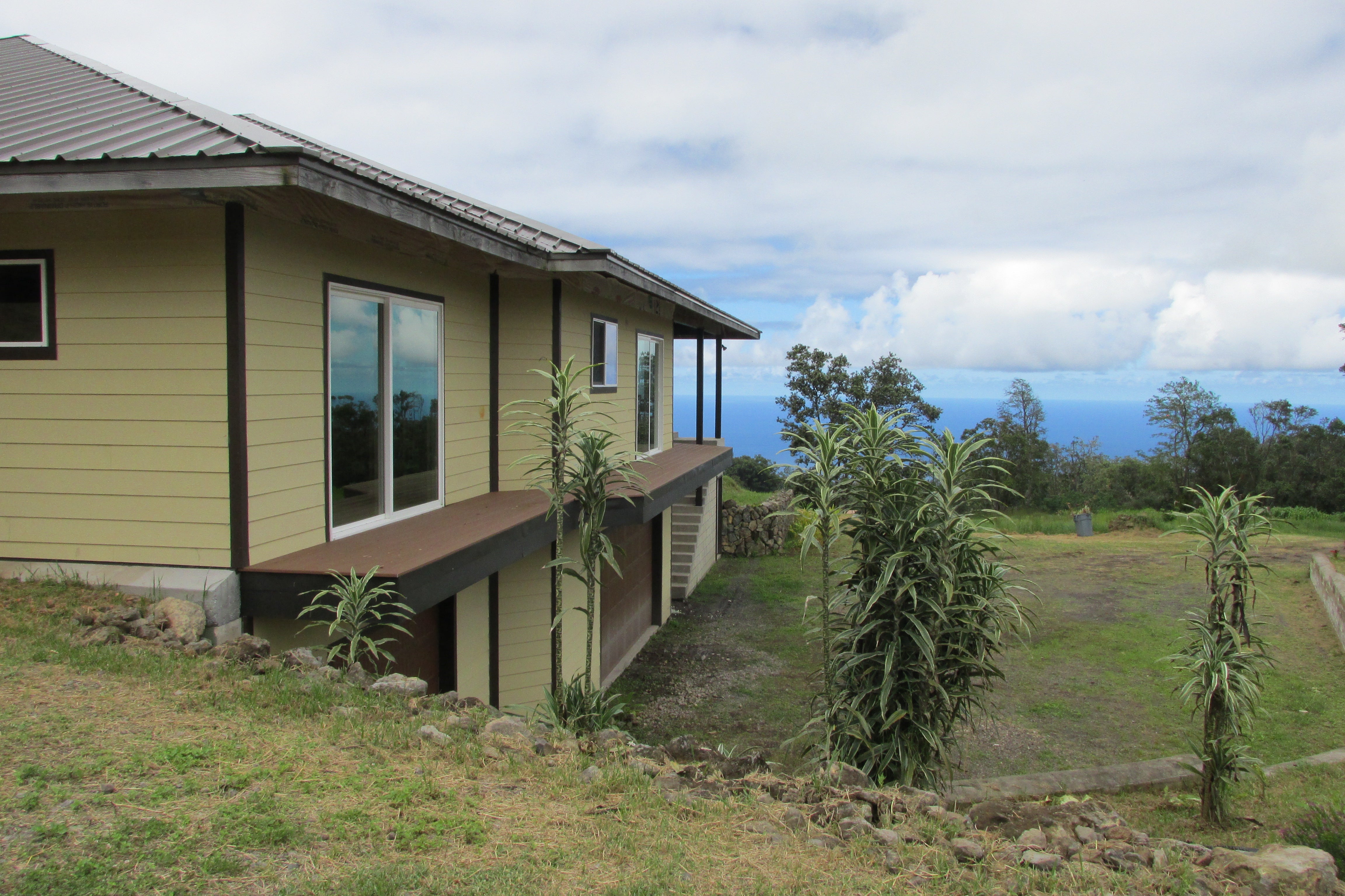 Pale yellow home with a raised deck overlooking a grassy slope and the ocean.
