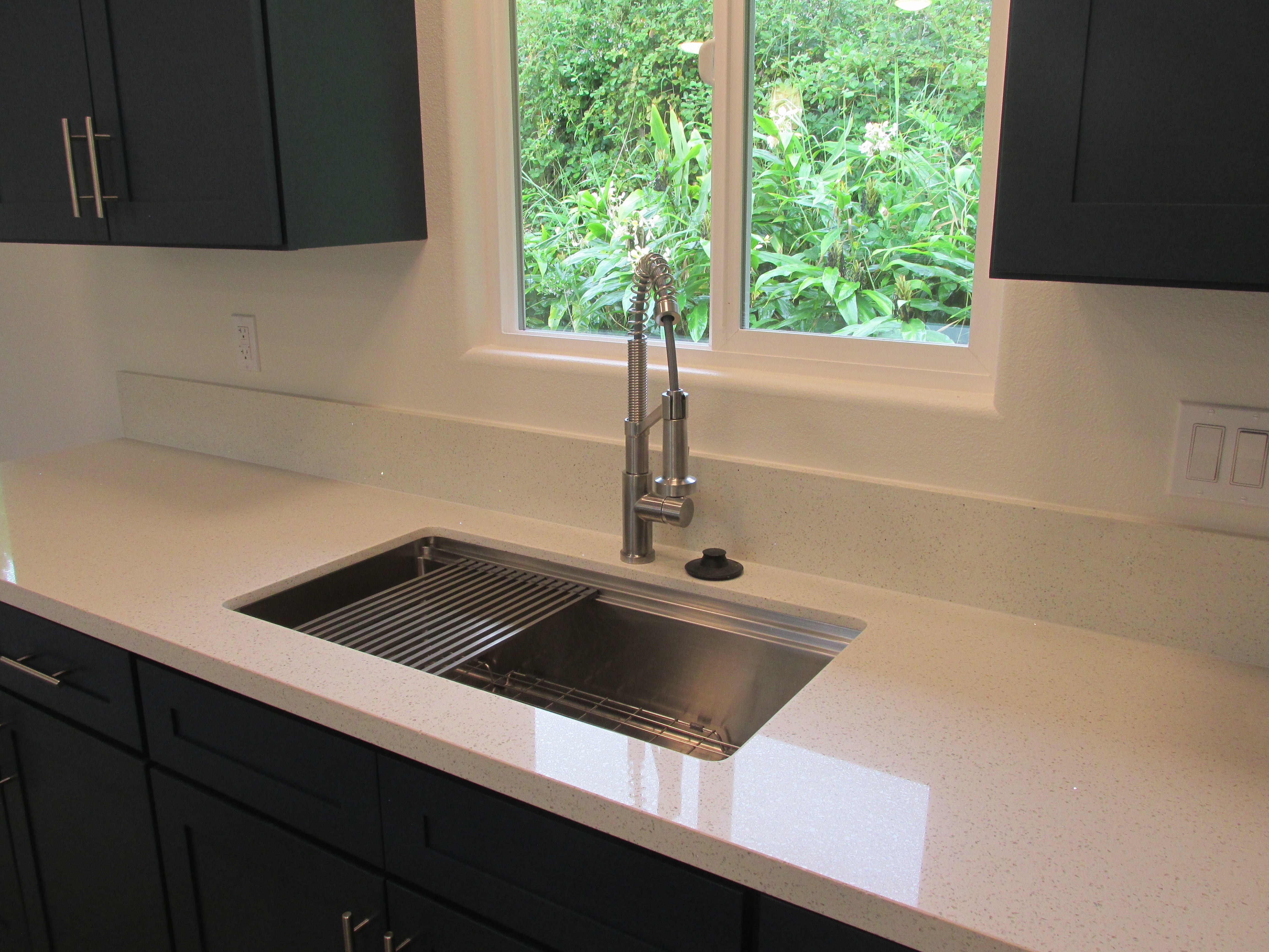 Kitchen sink with a spring-coil faucet, dark cabinets, and a window facing dense greenery.