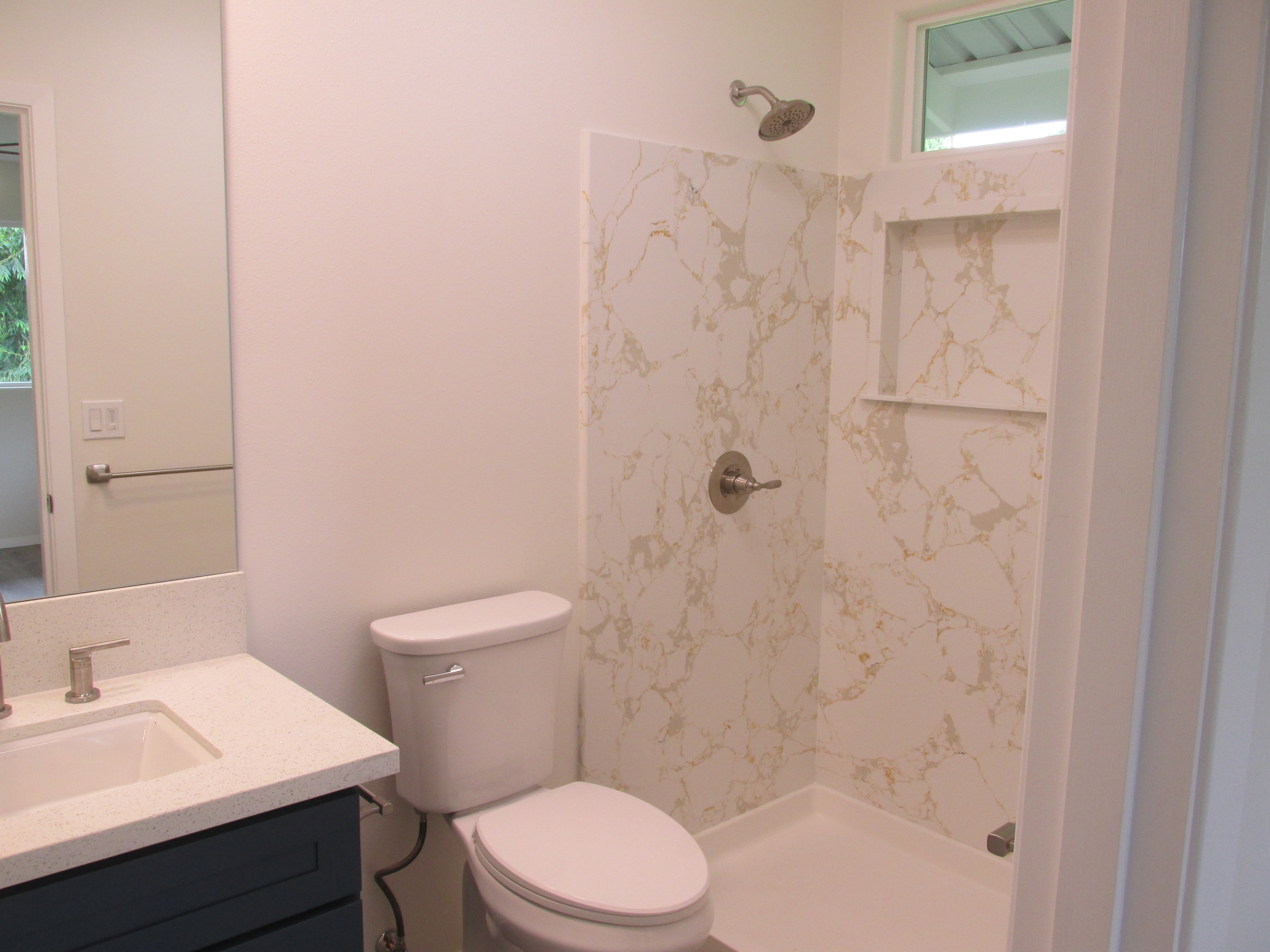 Bathroom with a marble-look shower, high window, white vanity, and dark blue cabinet.