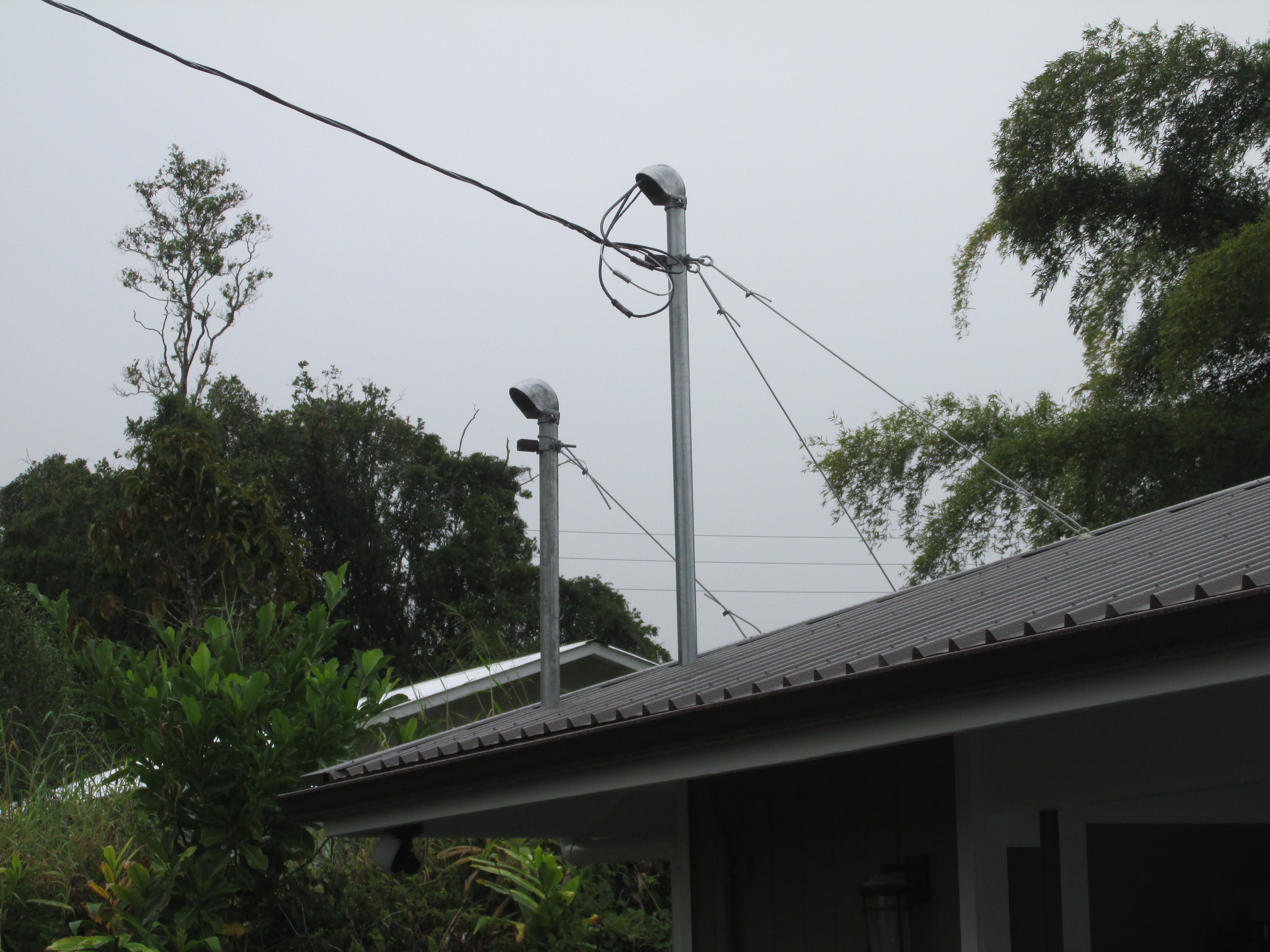 House roof with two metal vent pipes and utility wires amid dense tropical greenery.