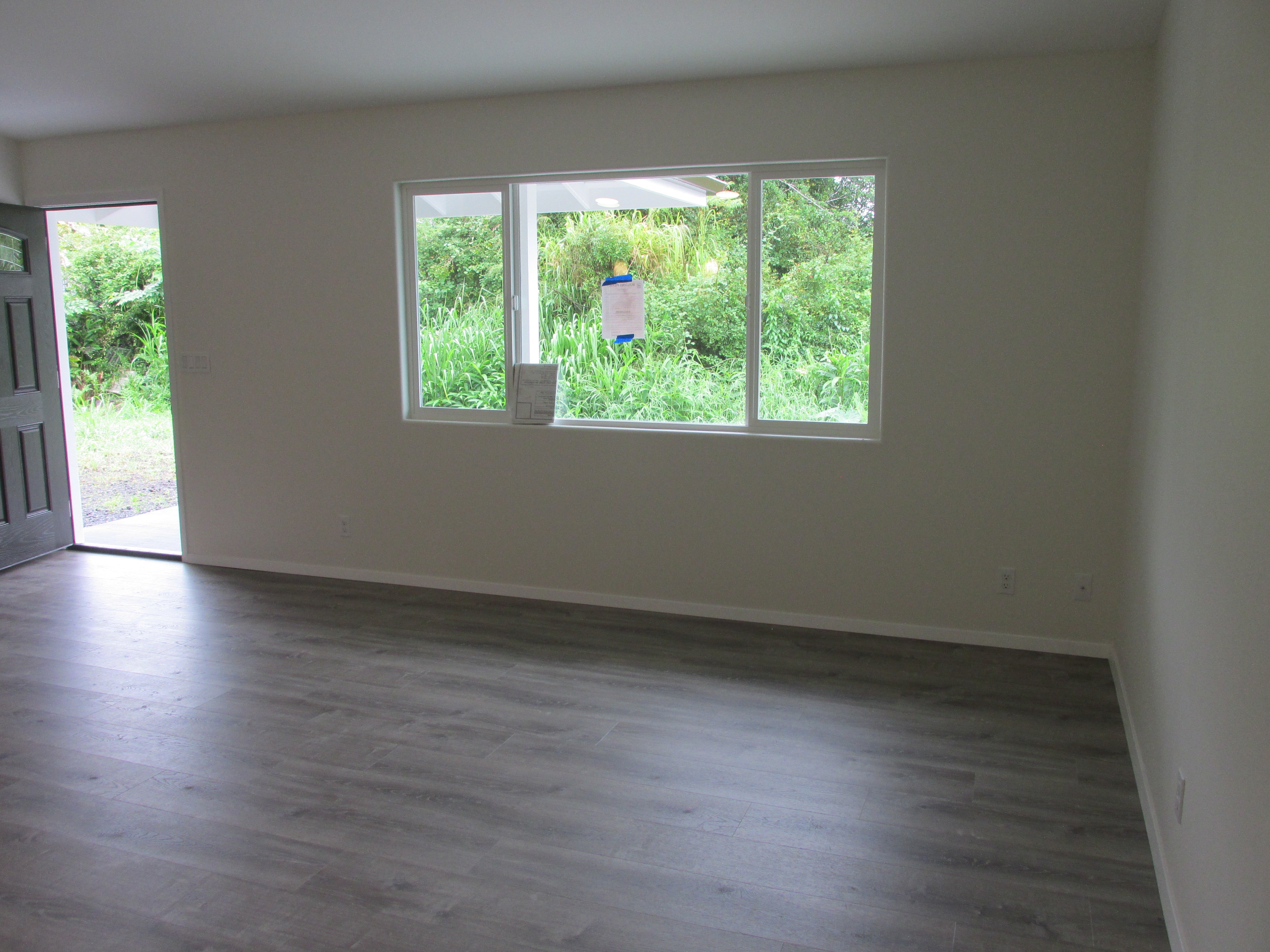 Vacant room with gray flooring, a large window overlooking greenery, and an open front door.
