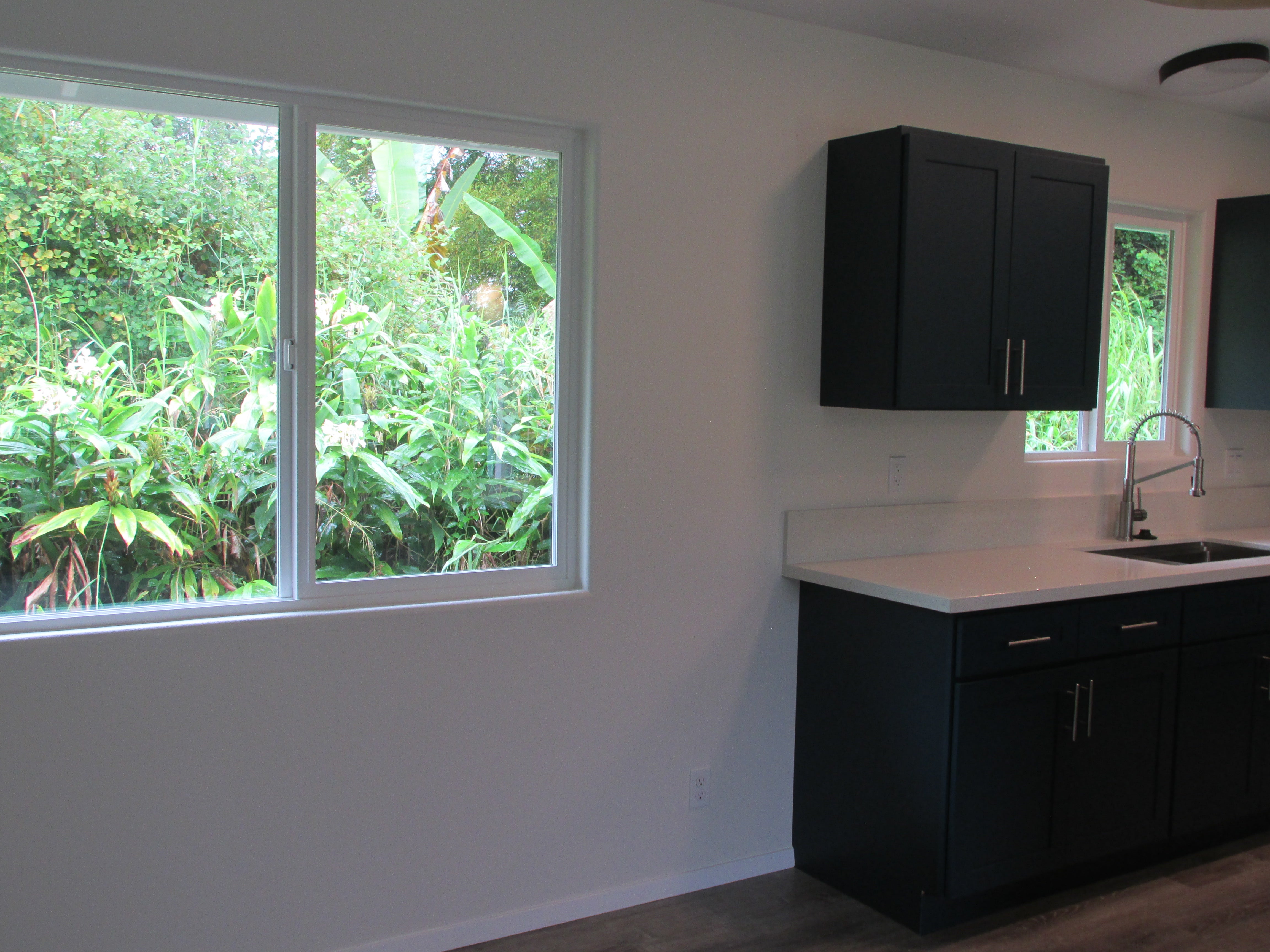 Kitchen with dark cabinets and white counters beside windows overlooking lush tropical greenery.
