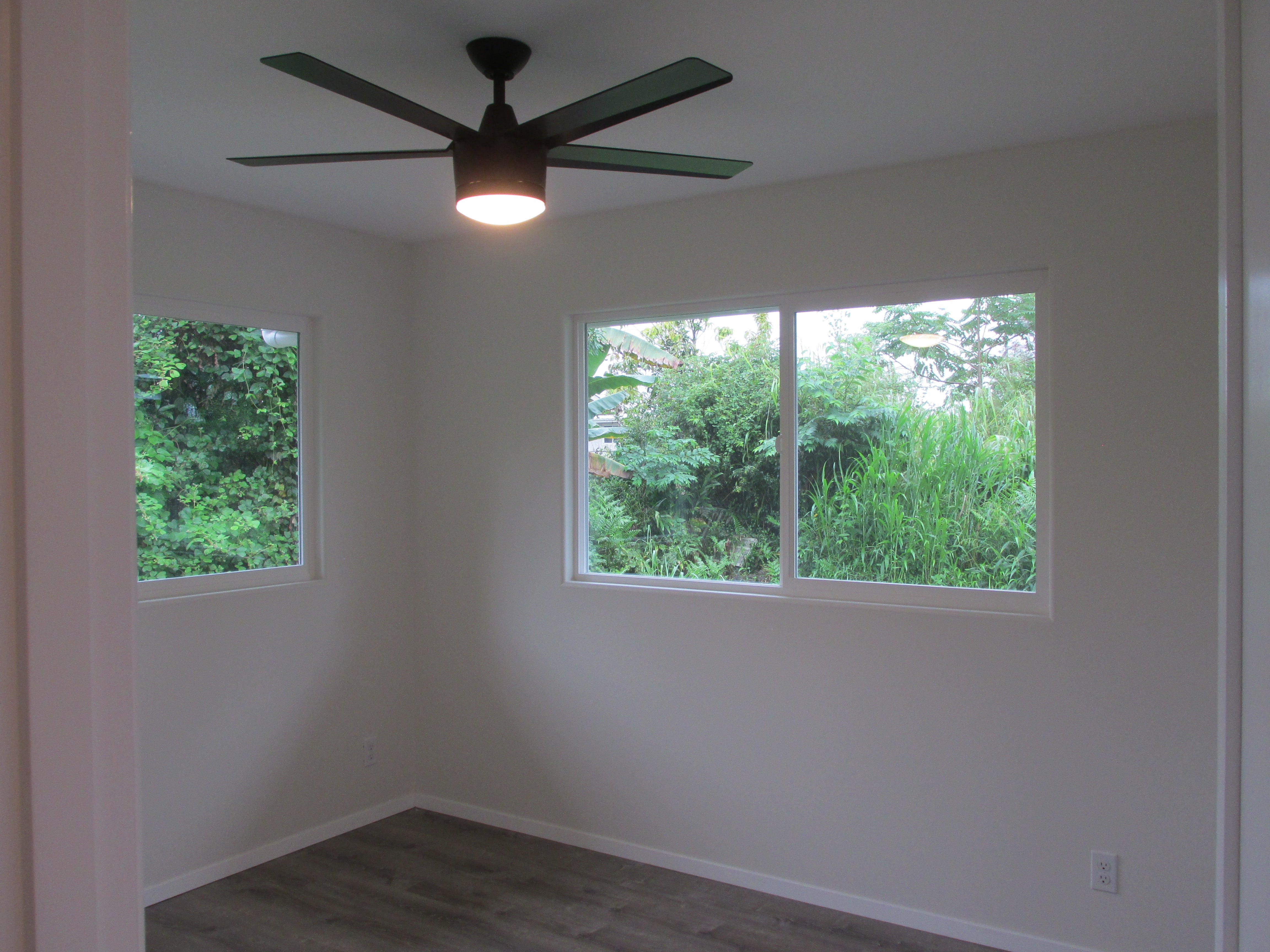 Empty room with a ceiling fan and windows overlooking dense tropical greenery.