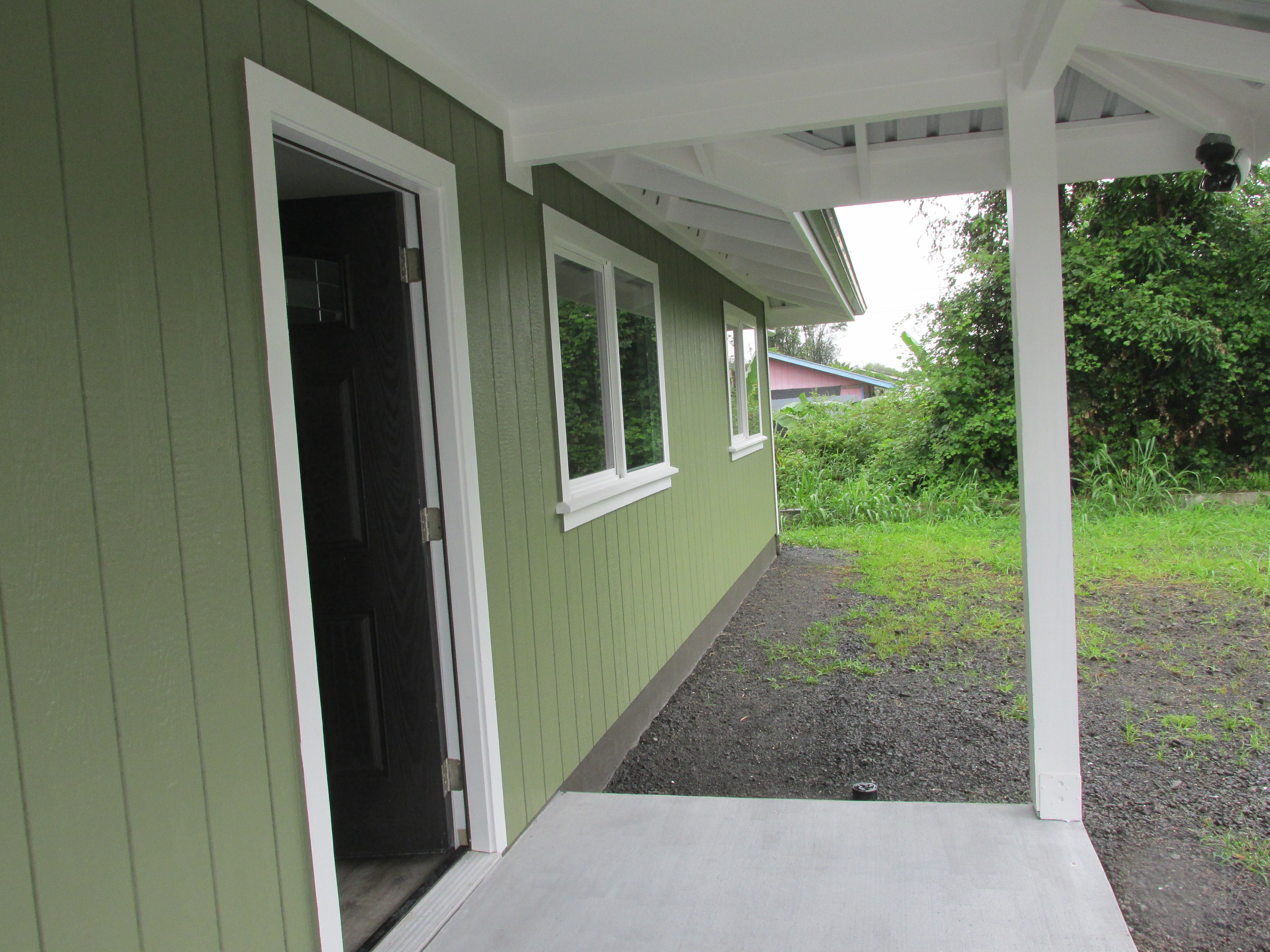 Covered porch beside an olive-green house, with an open doorway and white-trimmed windows.