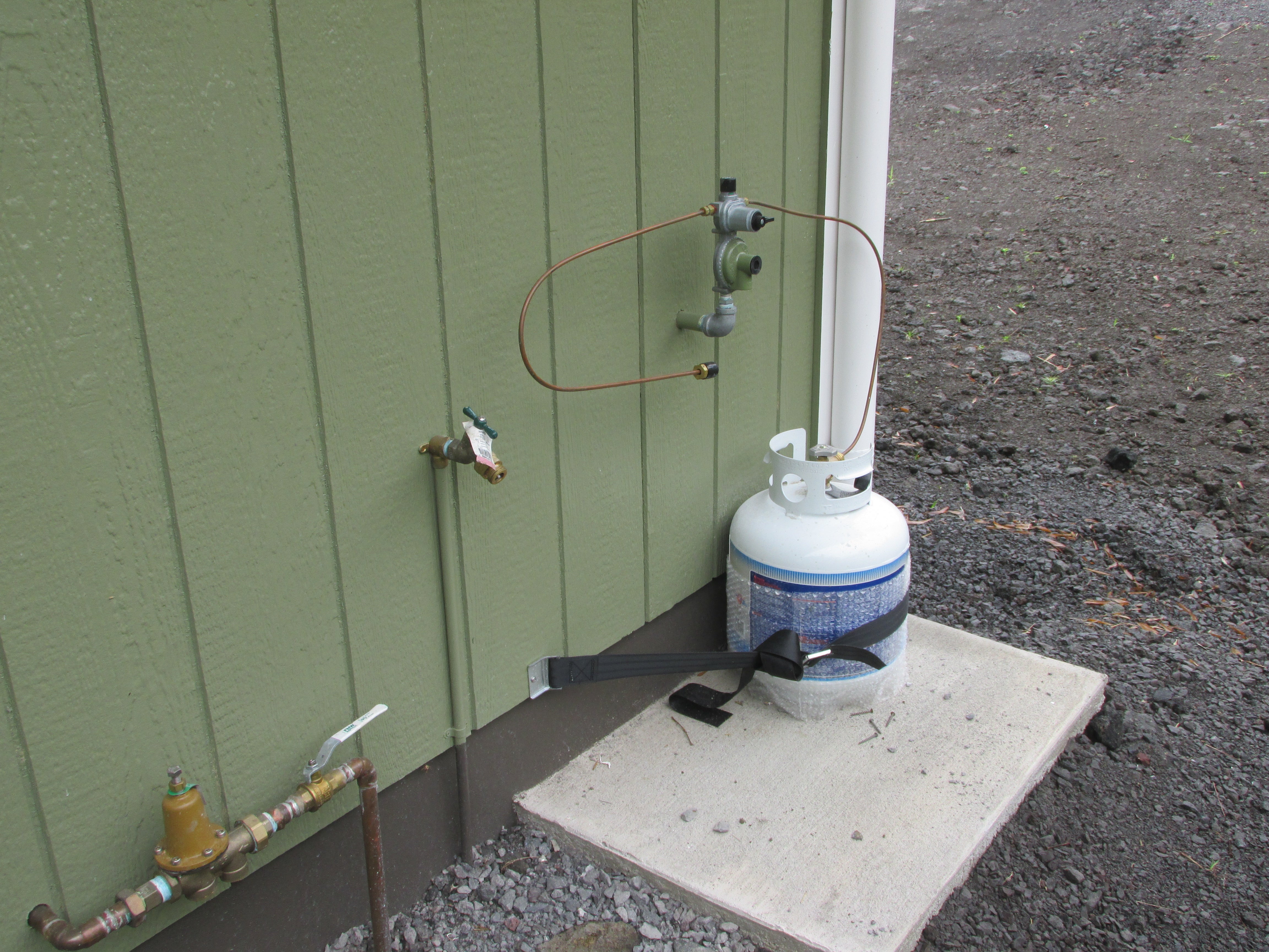 Exterior propane tank and gas connections beside a green-sided home.