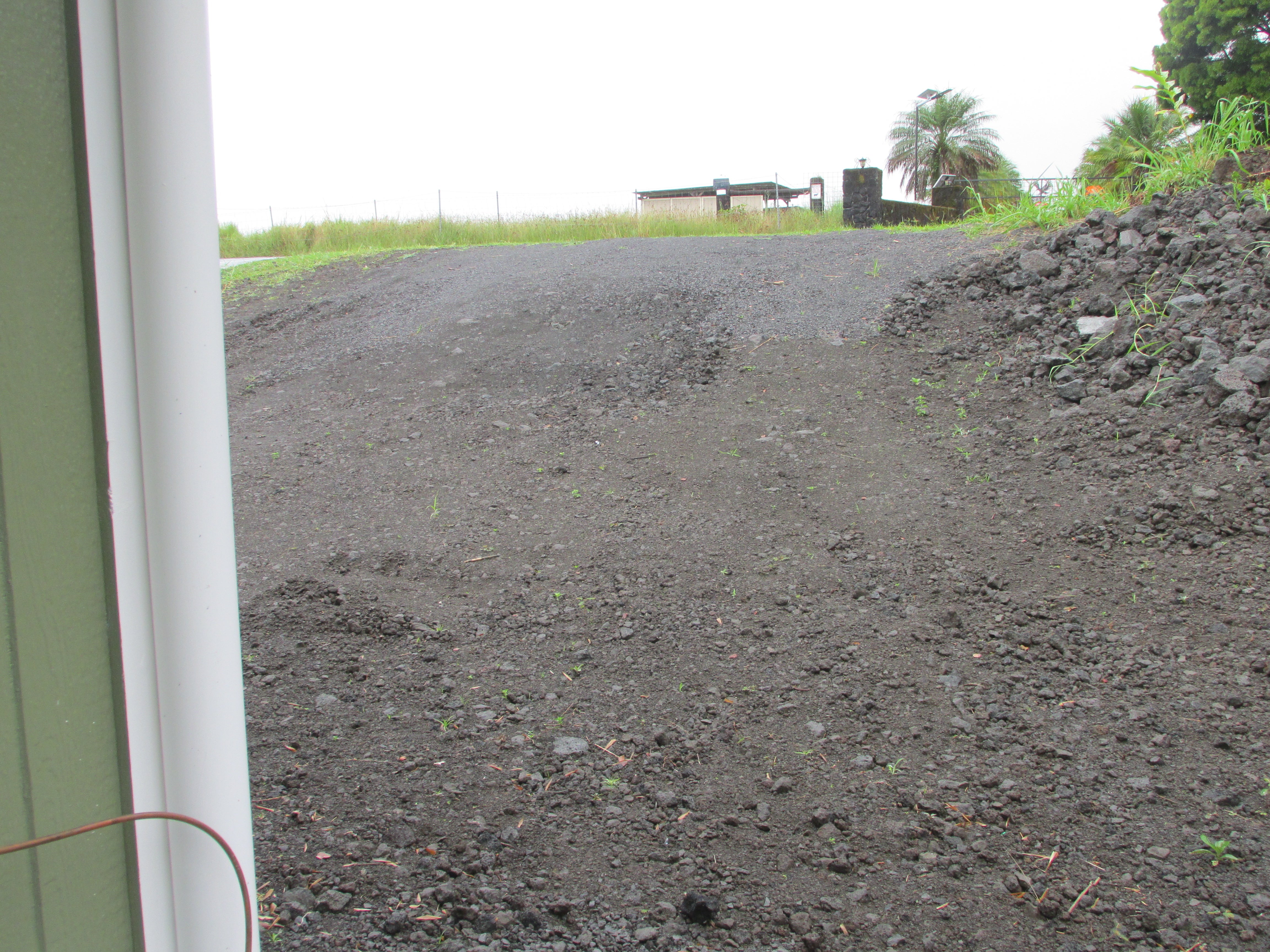 Gravel lot bordered by volcanic rock and tropical vegetation, with a fence and small structure in the distance.